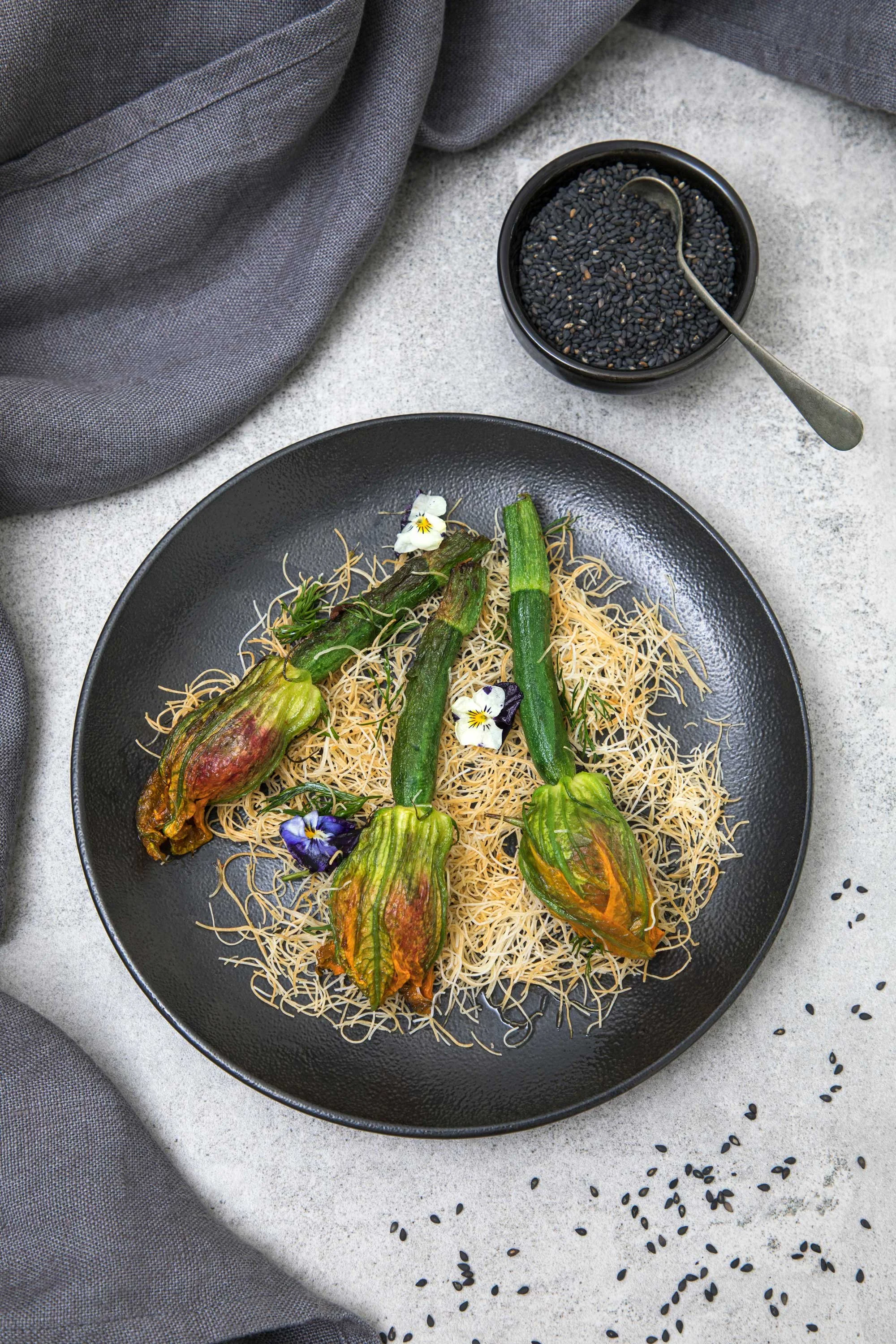 A black plate with stuffed squash blossoms on a bed of shredded cheese or pasta, garnished with edible flowers. A small black bowl of black sesame seeds with a silver spoon is nearby, with loose sesame seeds scattered on the light-colored surface, pa