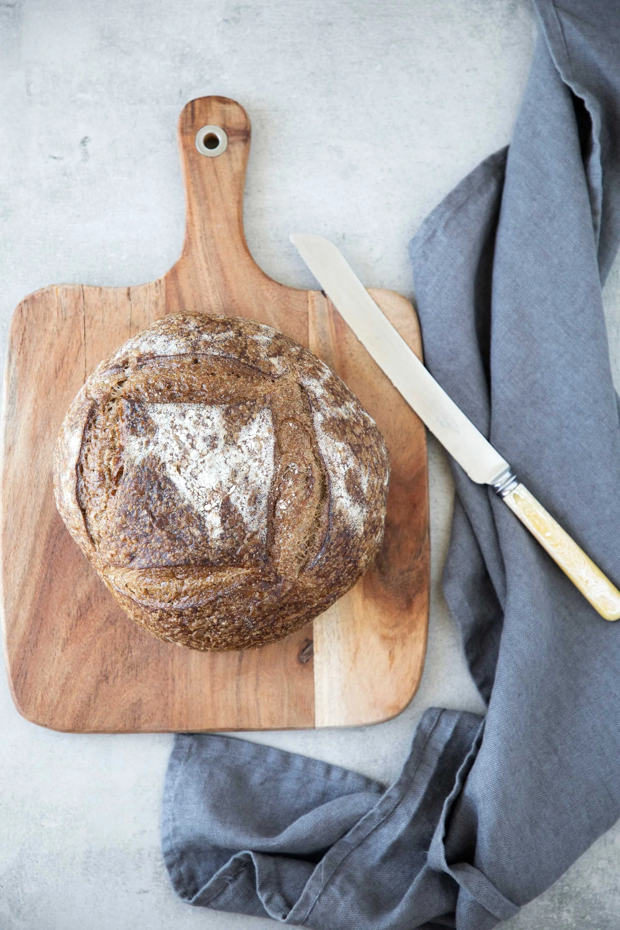 A round loaf of rustic bread with a browned crust and dusted with flour, resting on a wooden cutting board. A serrated bread knife with a cream-colored handle and a gray cloth are also on the board.