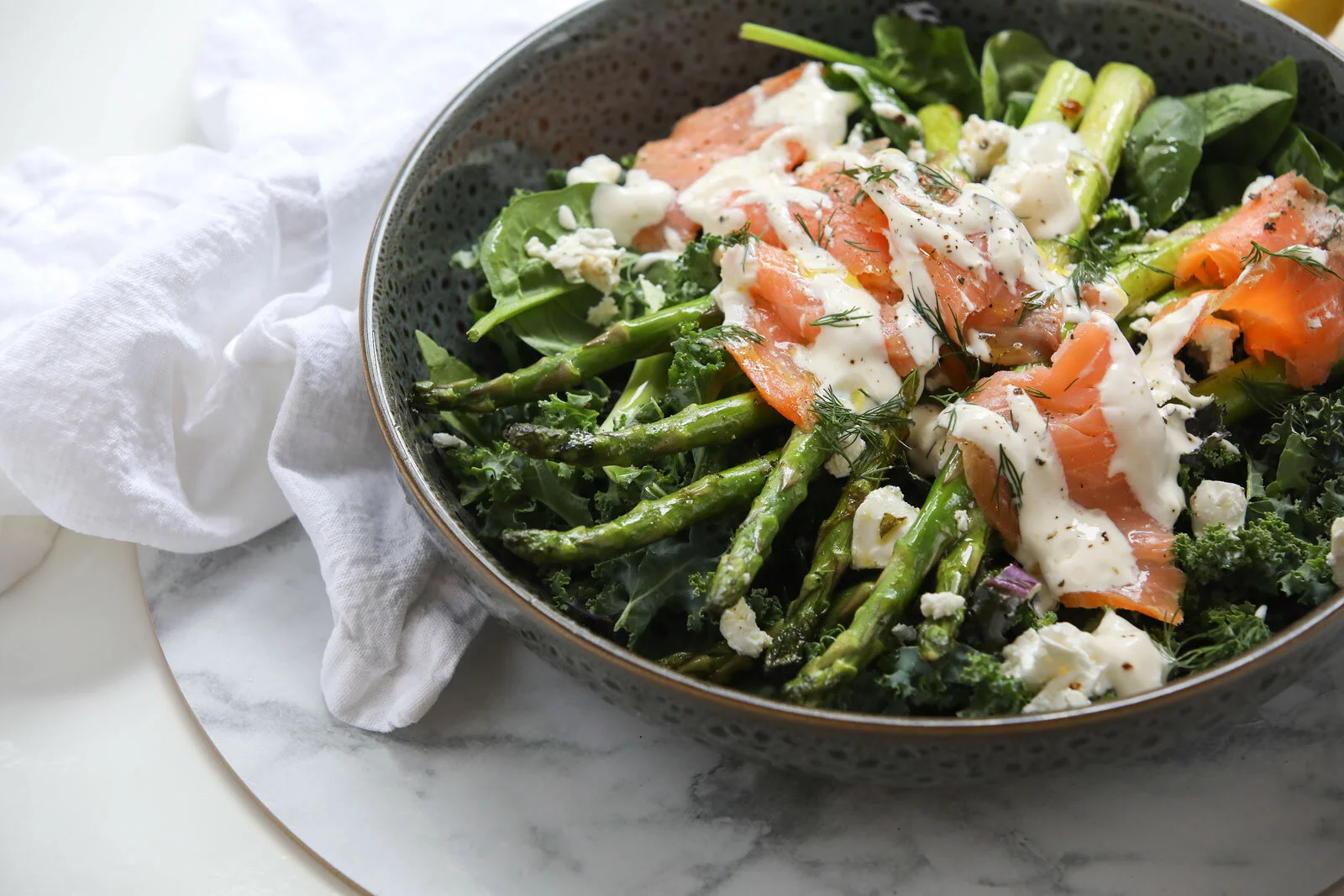 Salad with roasted asparagus, smoked salmon, leafy greens, crumbled cheese, and dill in a dark bowl on a white marble surface.