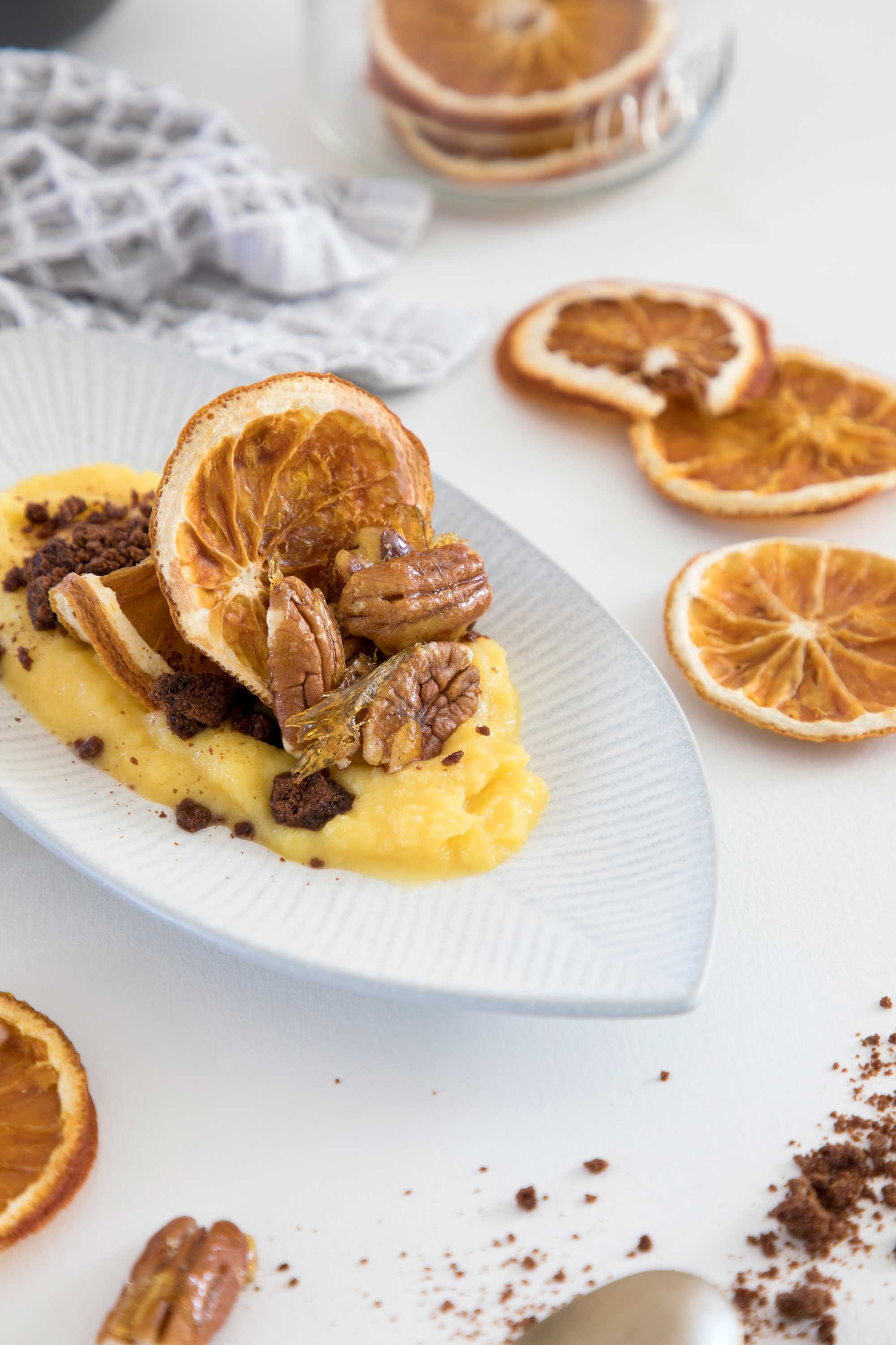 Close-up of dried orange slices on a white surface with scattered chocolate crumbs and pecans, with a plate of mashed sweet potatoes topped with dried orange slices and pecans in the background.