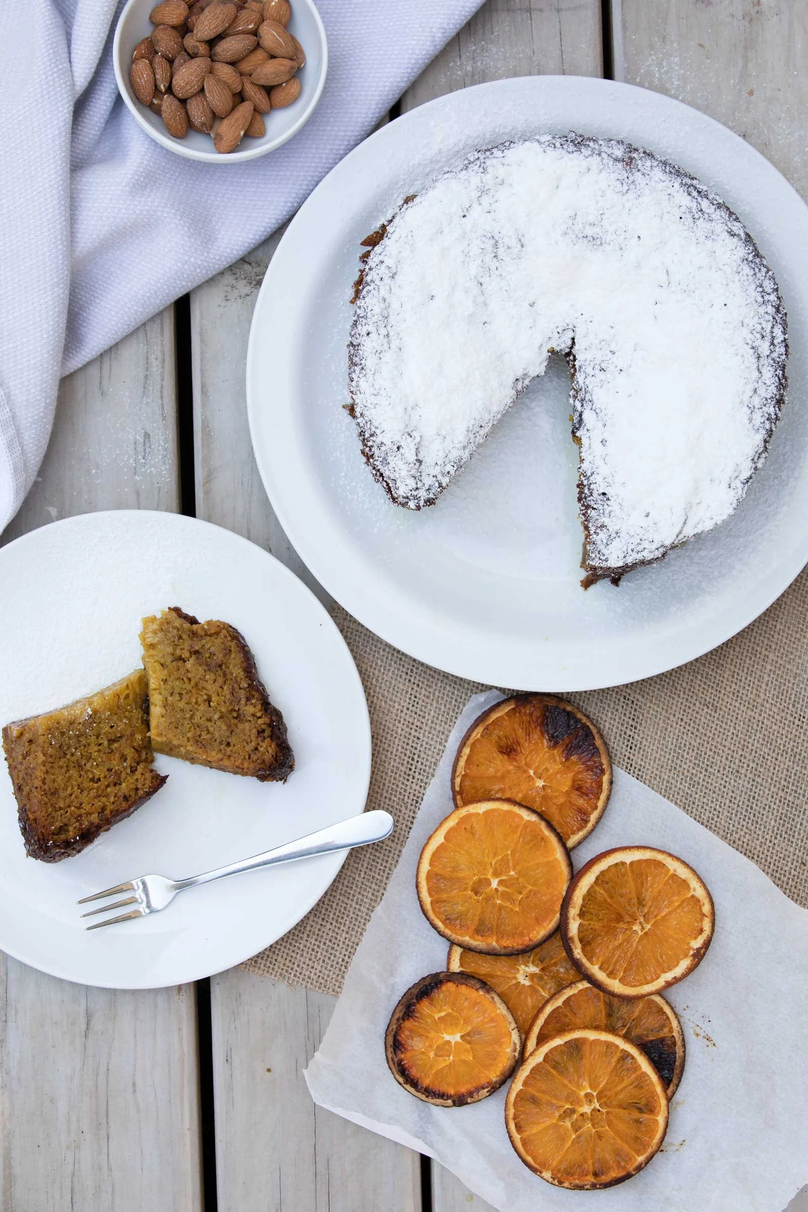 A white plate with a slice of fruitcake covered in powdered sugar, surrounded by slices of dried oranges, on a rustic wooden table.