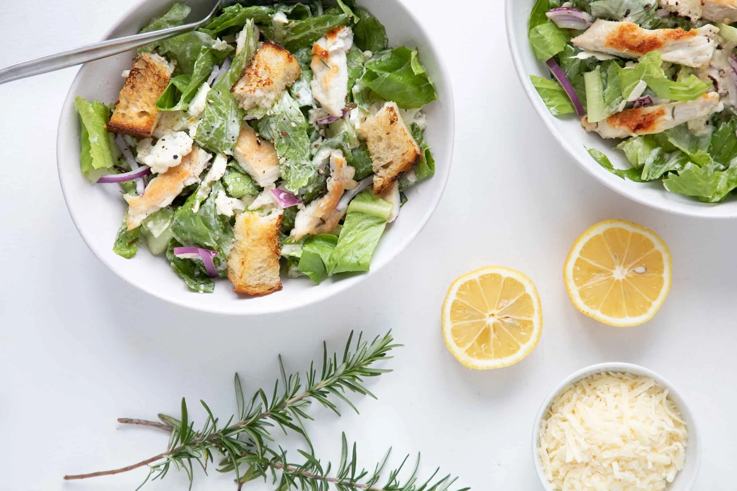 Close-up of two bowls of Caesar salad with croutons, shredded cheese, and lemon slices on a white surface.