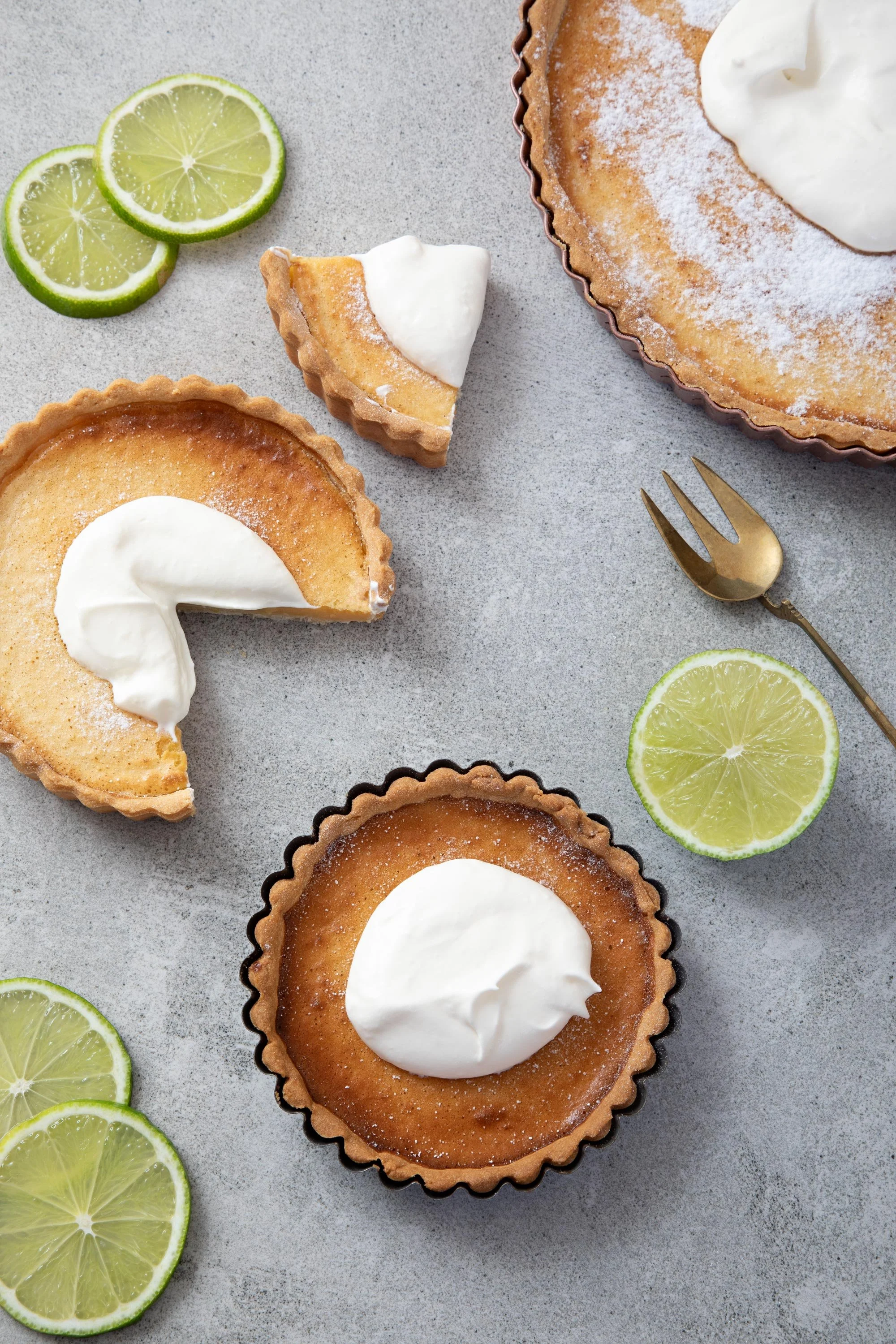 Lemon tart with a dollop of whipped cream in a tart mold, slices of fresh lime on a light gray surface.