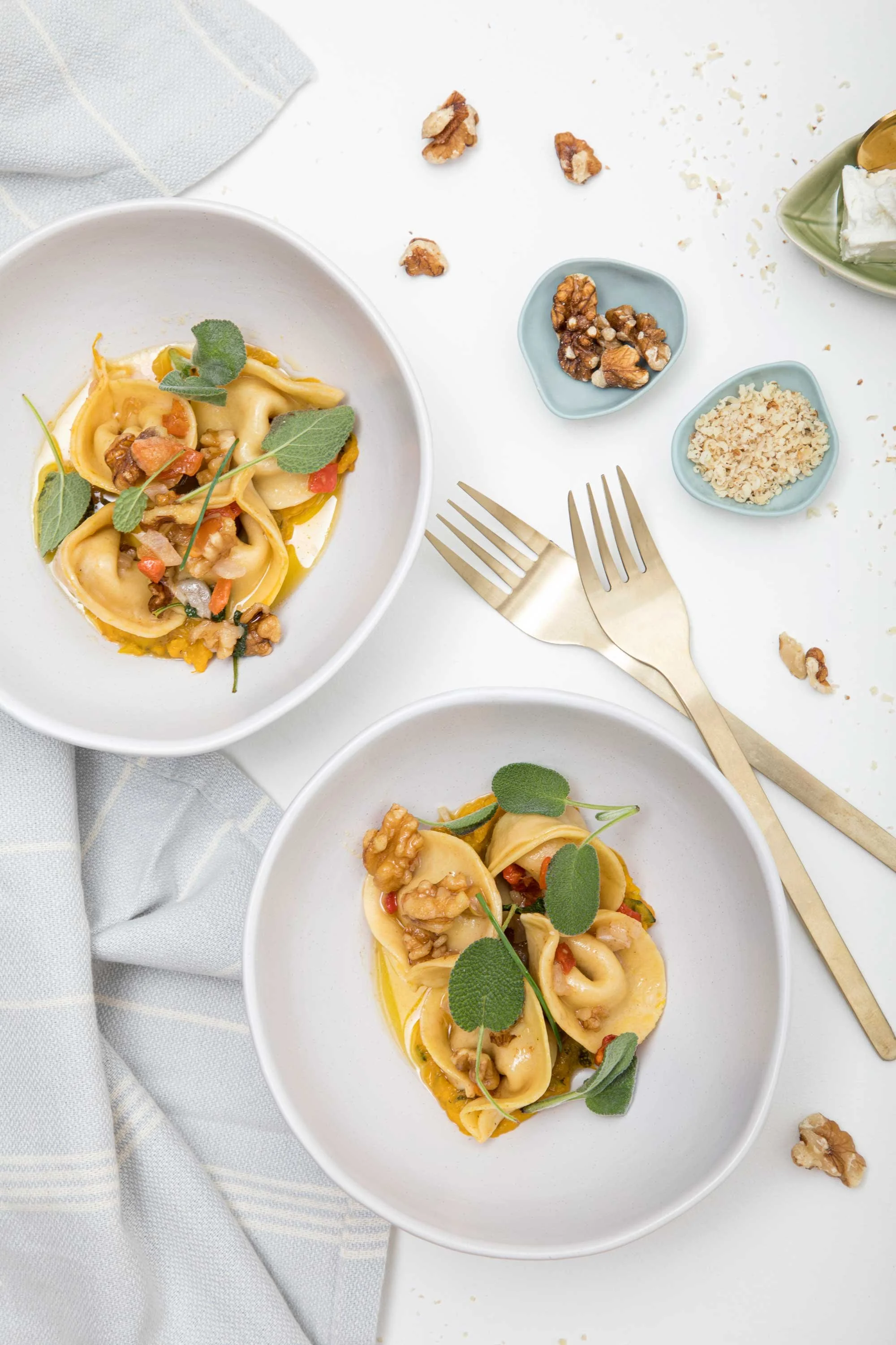 Close-up of two white bowls of tortellini pasta garnished with sage leaves, with small bowls of walnuts and breadcrumbs, silver forks, and scattered walnuts on a white tablecloth.