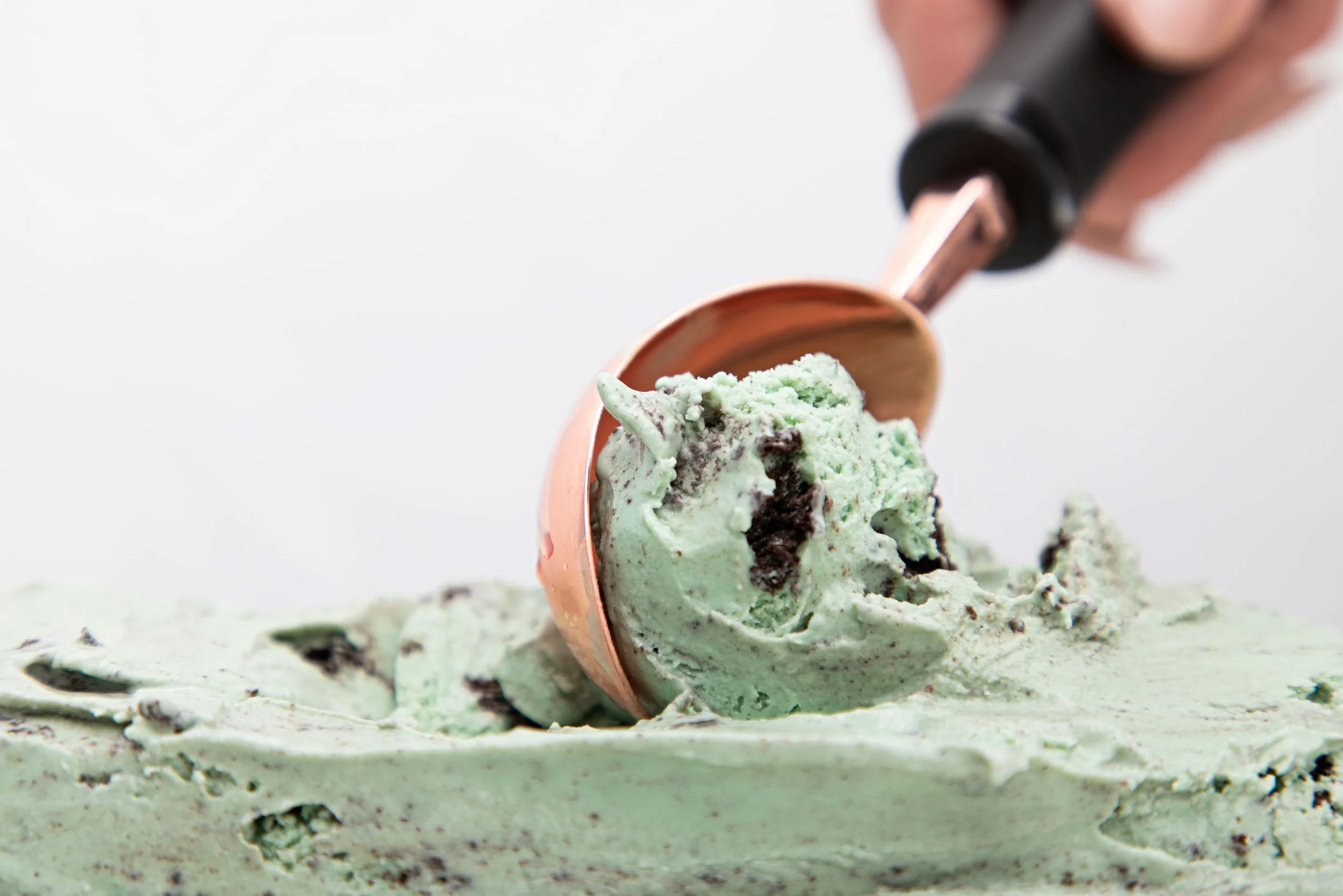 Mint chocolate chip ice cream in a copper cup with a black-handled ice cream scoop, melting slightly and sprinkled with chocolate chips.