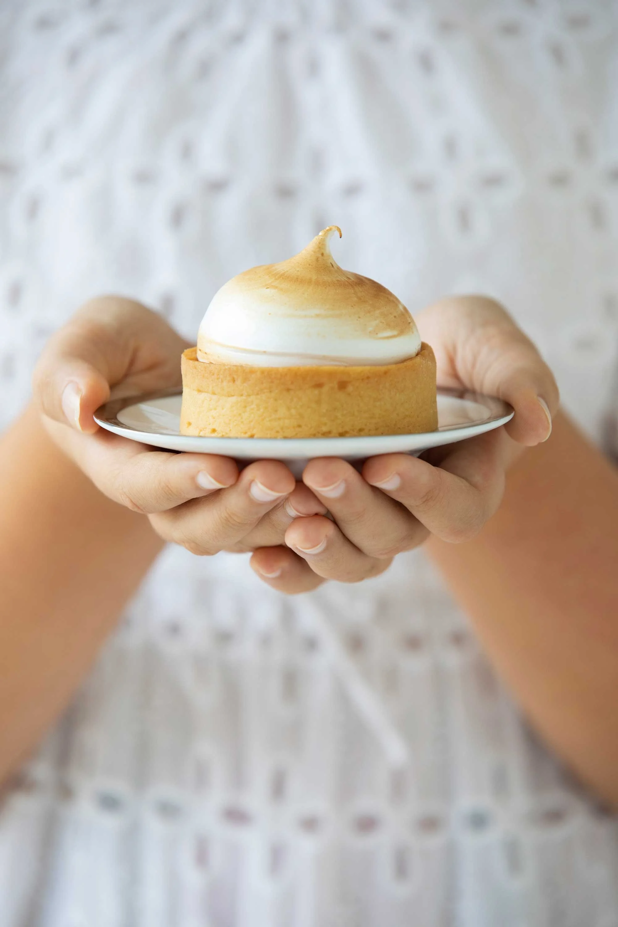 Person holding a white plate with a tart topped with meringue or whipped cream. The person wears a white shirt with a dotted pattern.
