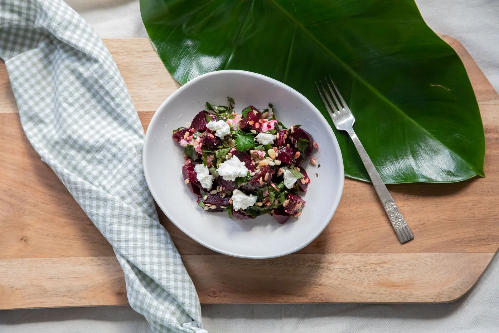 Bowl of beet salad with crumbled cheese, chopped herbs, and sunflower seeds on a wooden cutting board with a green cloth, a large leaf, and a silver fork nearby.