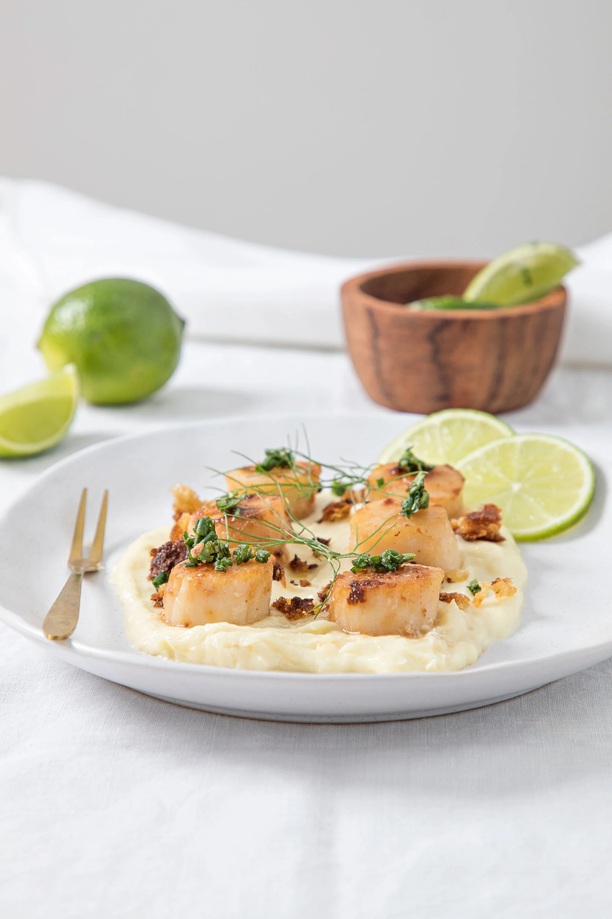 A plate of shrimp and scallops served over creamy mashed potatoes with lemon wedges and herbs, with limes and a wooden bowl with lime slices in the background, on a white table with a white cloth.