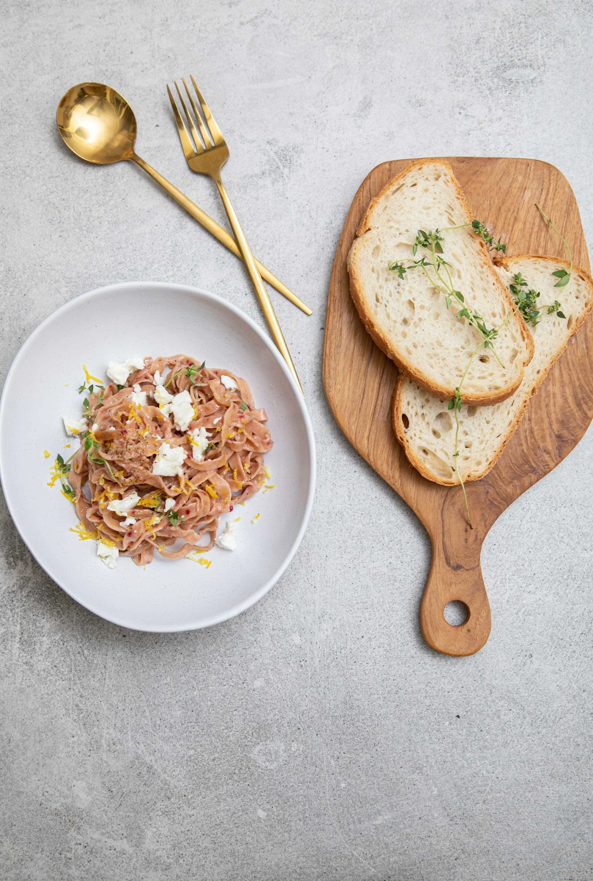 A bowl of pasta with a creamy sauce, garnished with crumbled cheese and herbs, along with bread slices topped with herbs on a wooden cutting board, and two gold utensils on a light-colored surface.