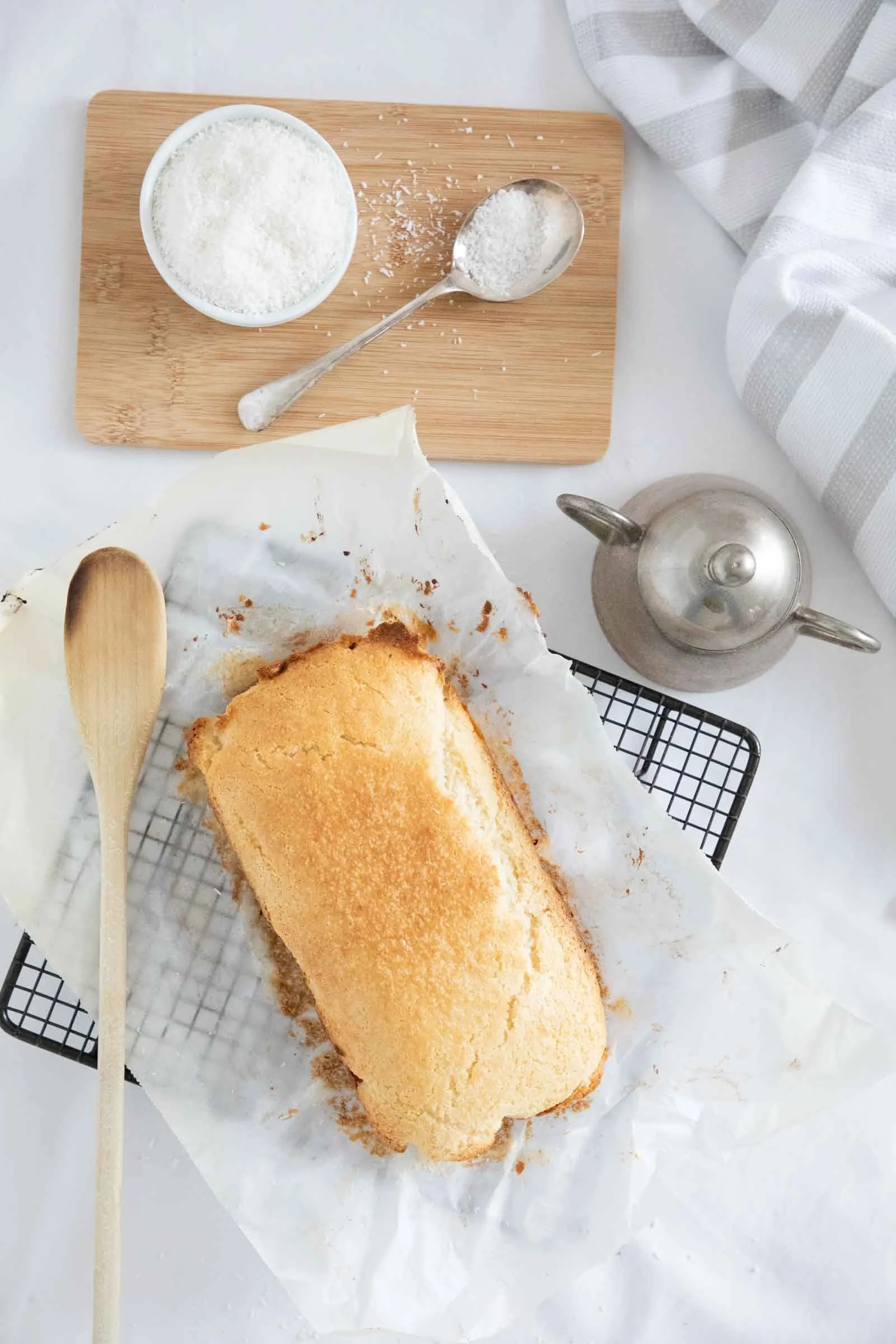 A partially eaten loaf of yellow cake on parchment paper, placed on a cooling rack, with a wooden spoon beside it. A bowl of shredded white coconut and a spoonful of coconut on a wooden board are in the background, along with a silver teapot on a whi