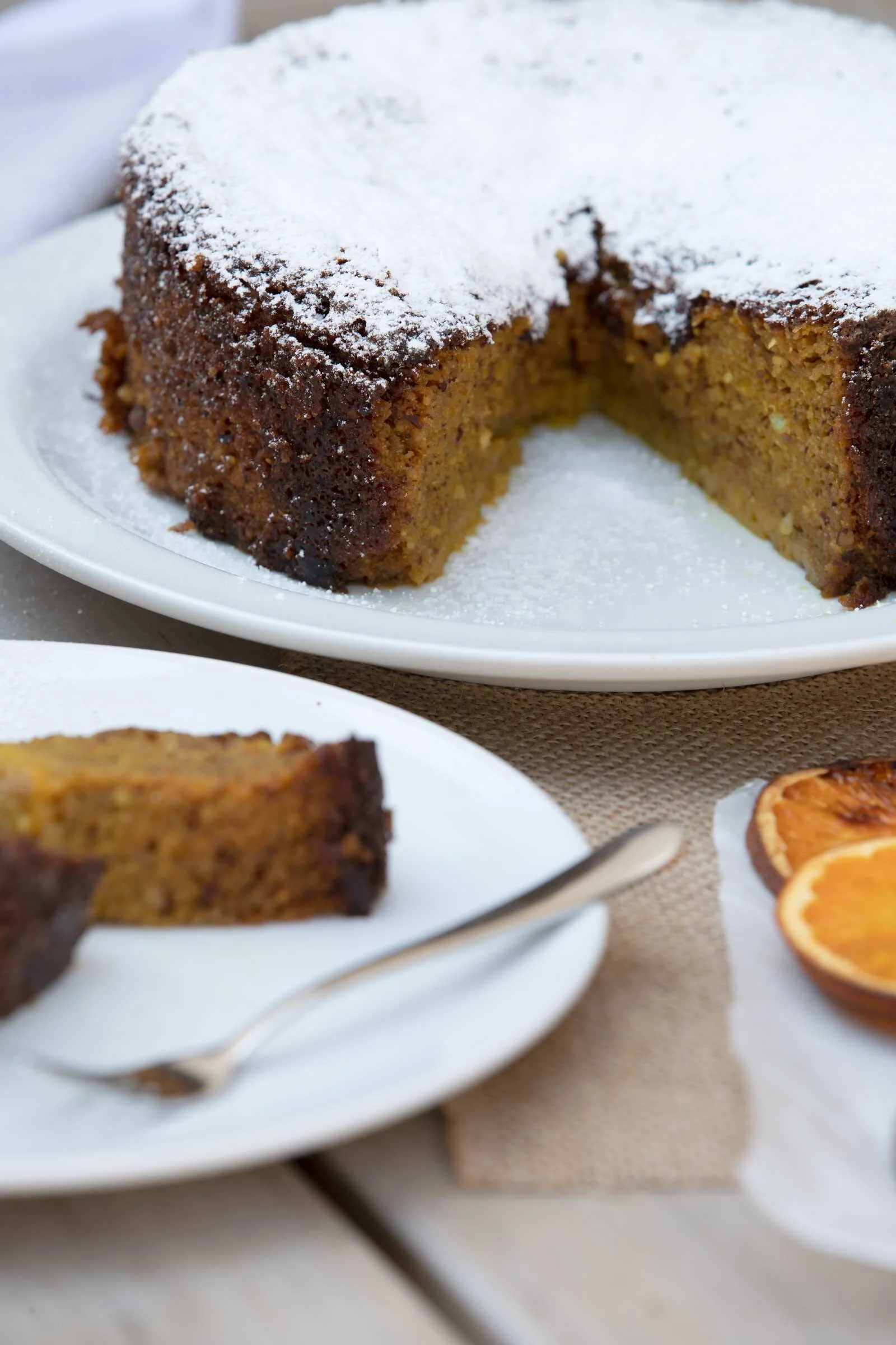 Close-up of a slice of pumpkin bread on a white plate with powdered sugar on top, with a partially visible piece in the background, and a small dish of sliced oranges to the side.