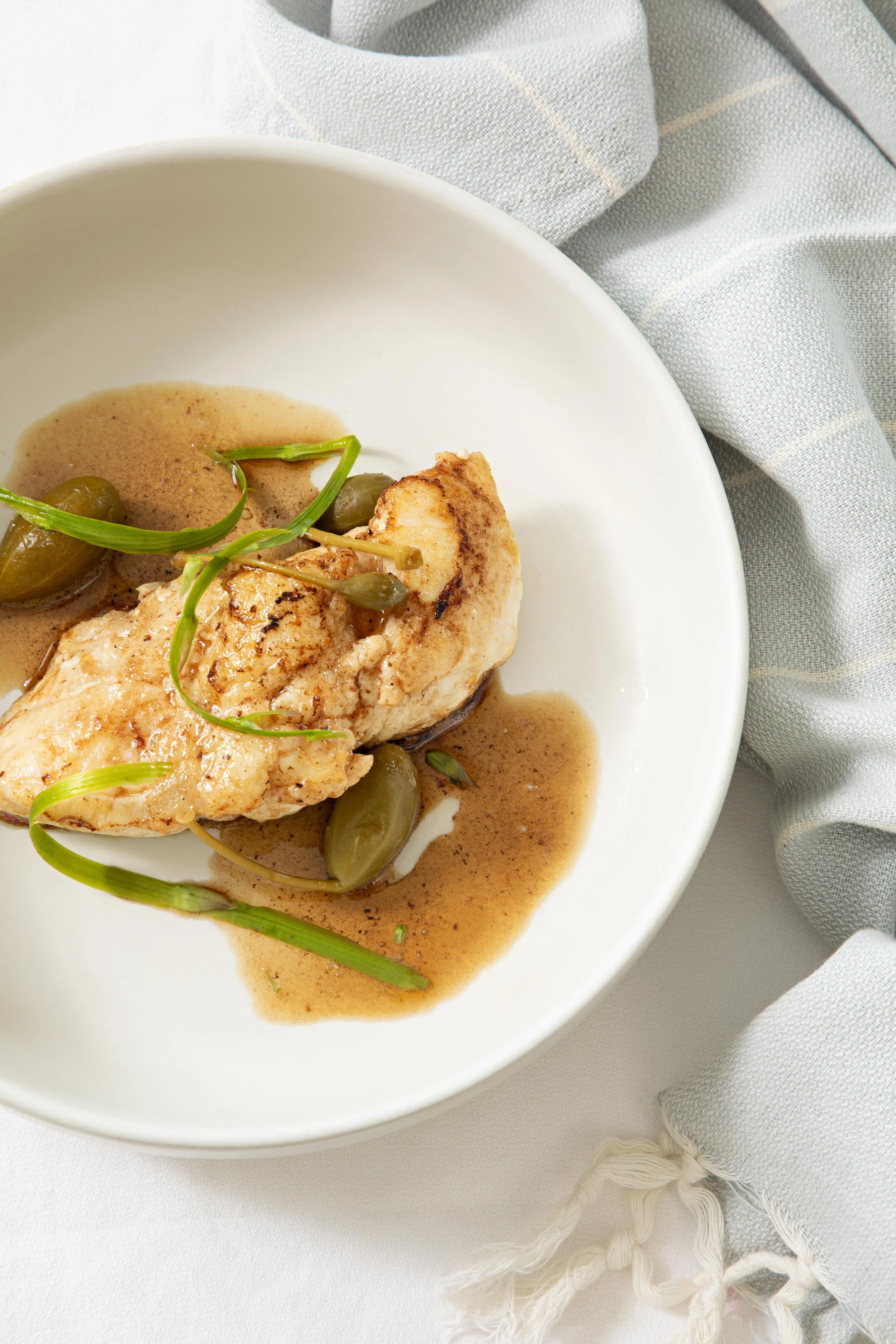 A white bowl contains a piece of grilled chicken topped with green microgreens and capers, served with a light brown sauce. The bowl is placed on a white surface with a light-colored, striped cloth nearby.