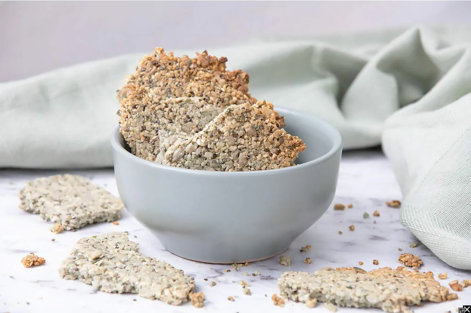 Granola bars in a gray bowl with some granola bars on the white surface around the bowl, and a light-colored cloth in the background.