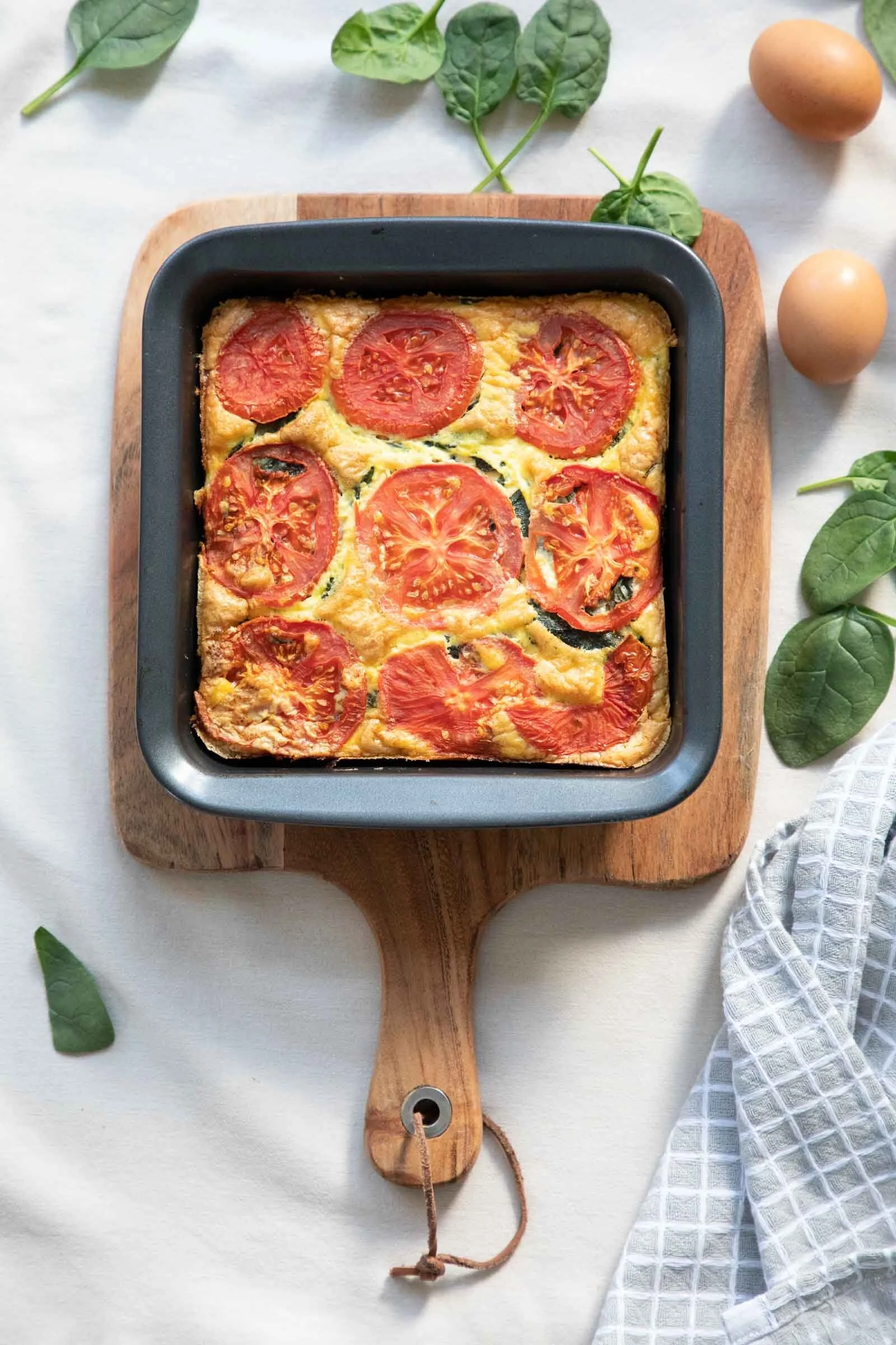Baking dish with tomato and egg casserole on wooden cutting board, surrounded by fresh spinach leaves, two brown eggs, and a white checkered cloth on a white surface.