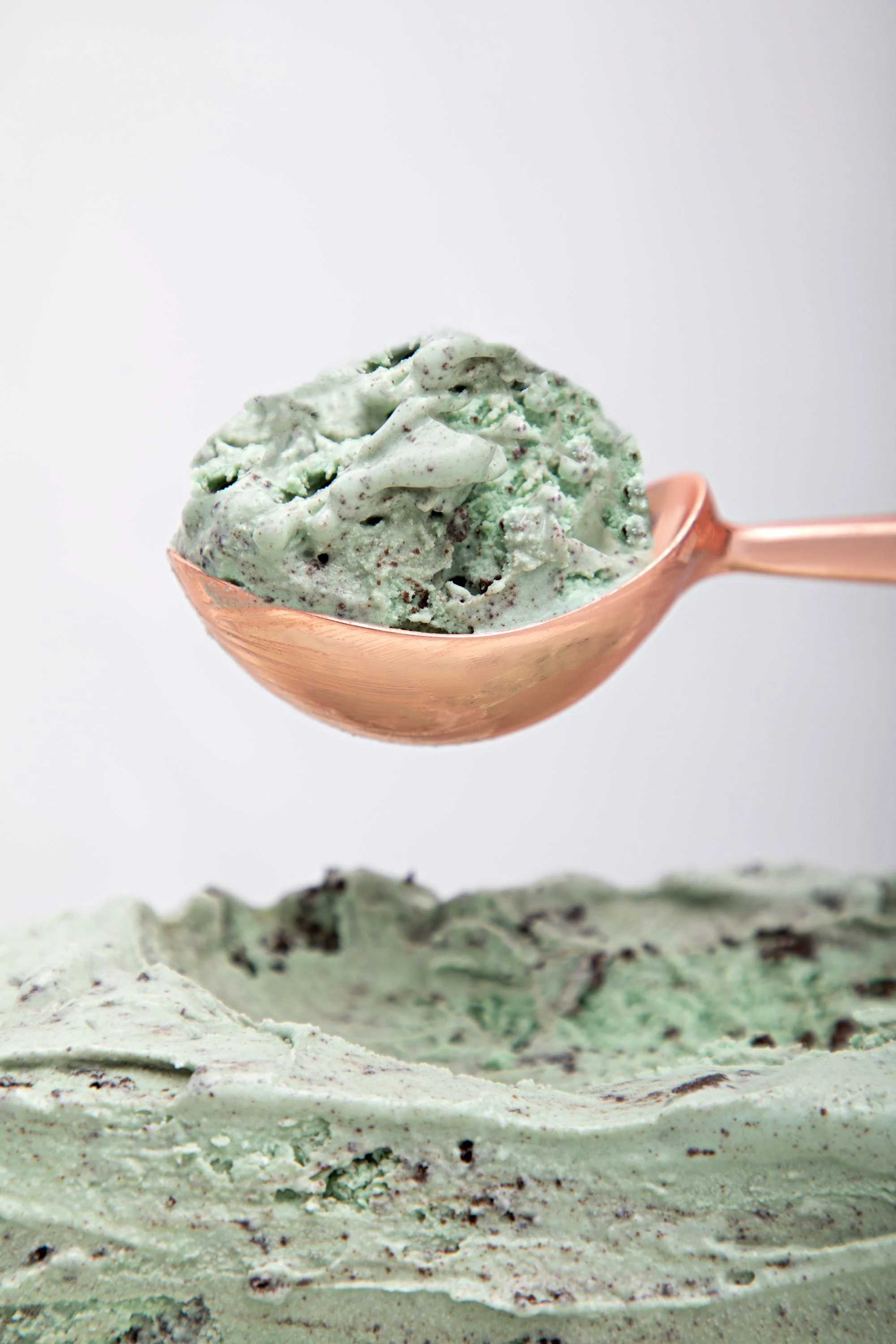 Close-up of a scoop of mint chocolate chip ice cream in a copper bowl, with a textured mint ice cream base and chocolate pieces, against a plain light background.