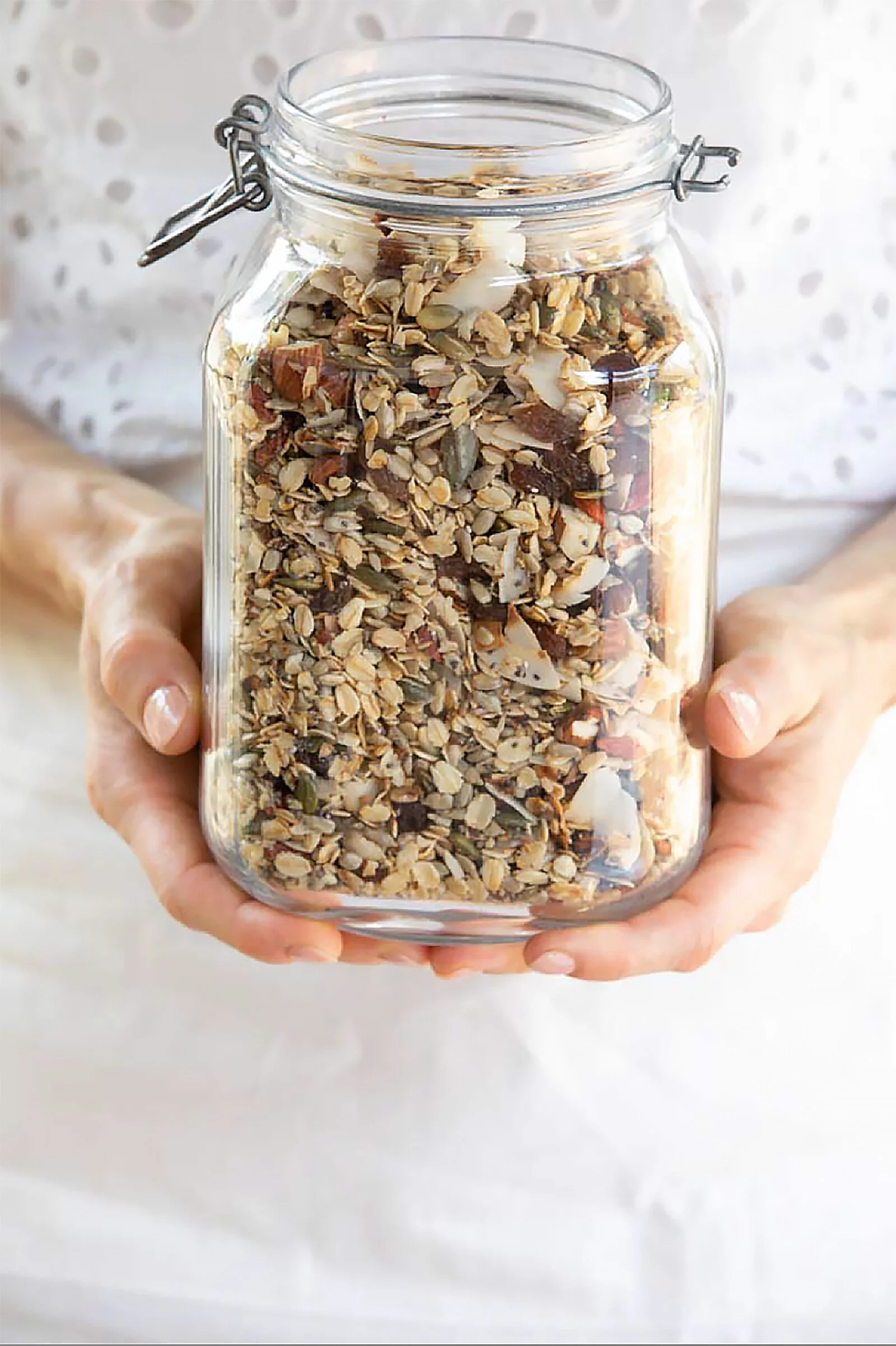 A person holding a glass jar filled with homemade trail mix containing various nuts, seeds, and dried fruits.