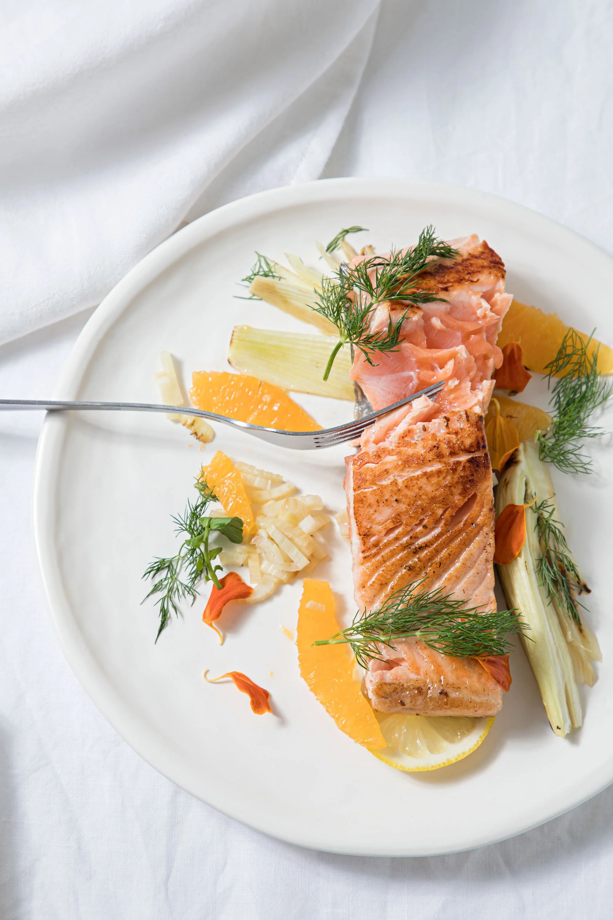 A plate with a cooked salmon fillet garnished with fresh dill, served with orange, lemon, and pineapple slices, and accompanied by roasted vegetables, on a white tablecloth.
