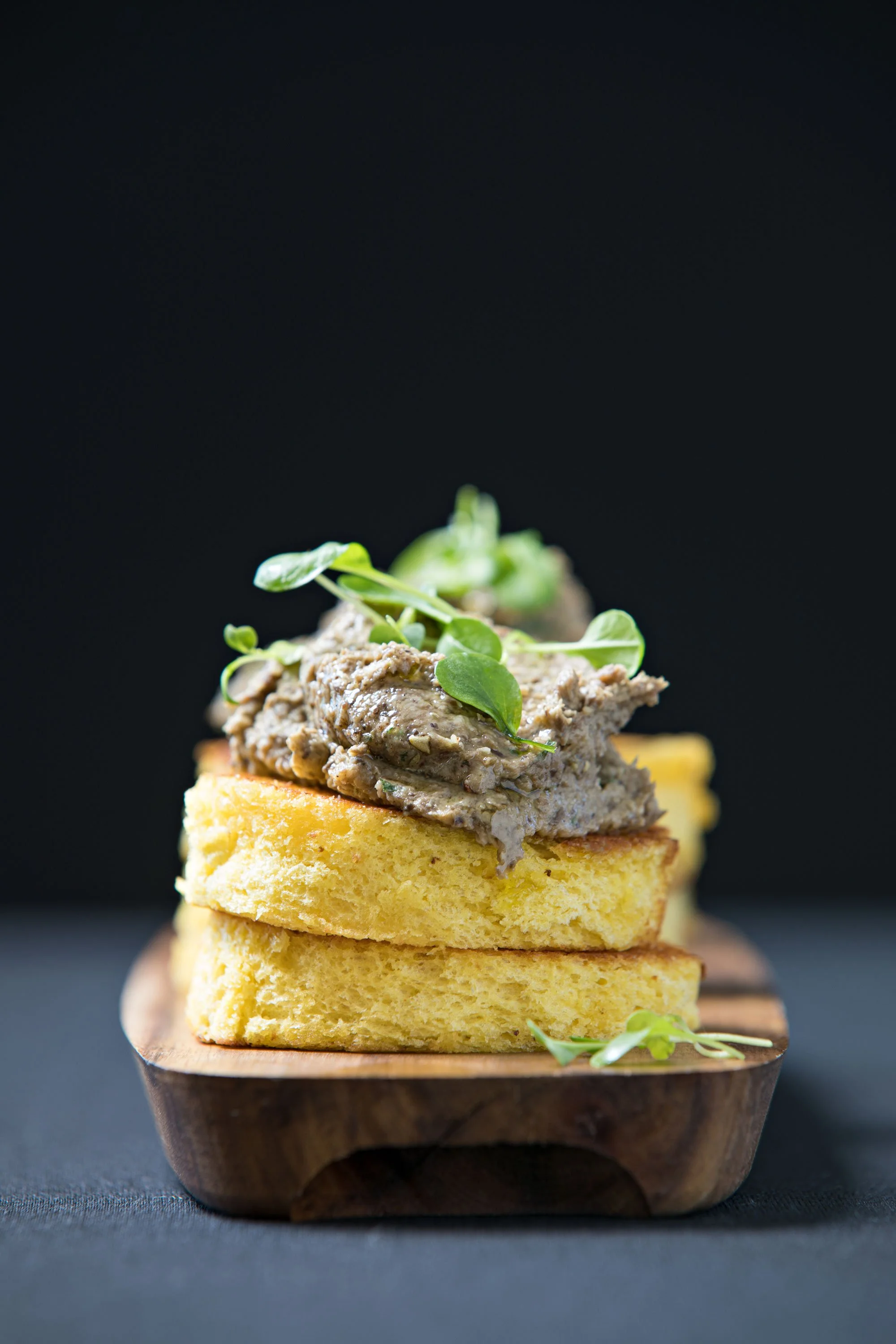Stacked slices of yellow bread topped with a meat spread and garnished with microgreens on a wooden serving board.
