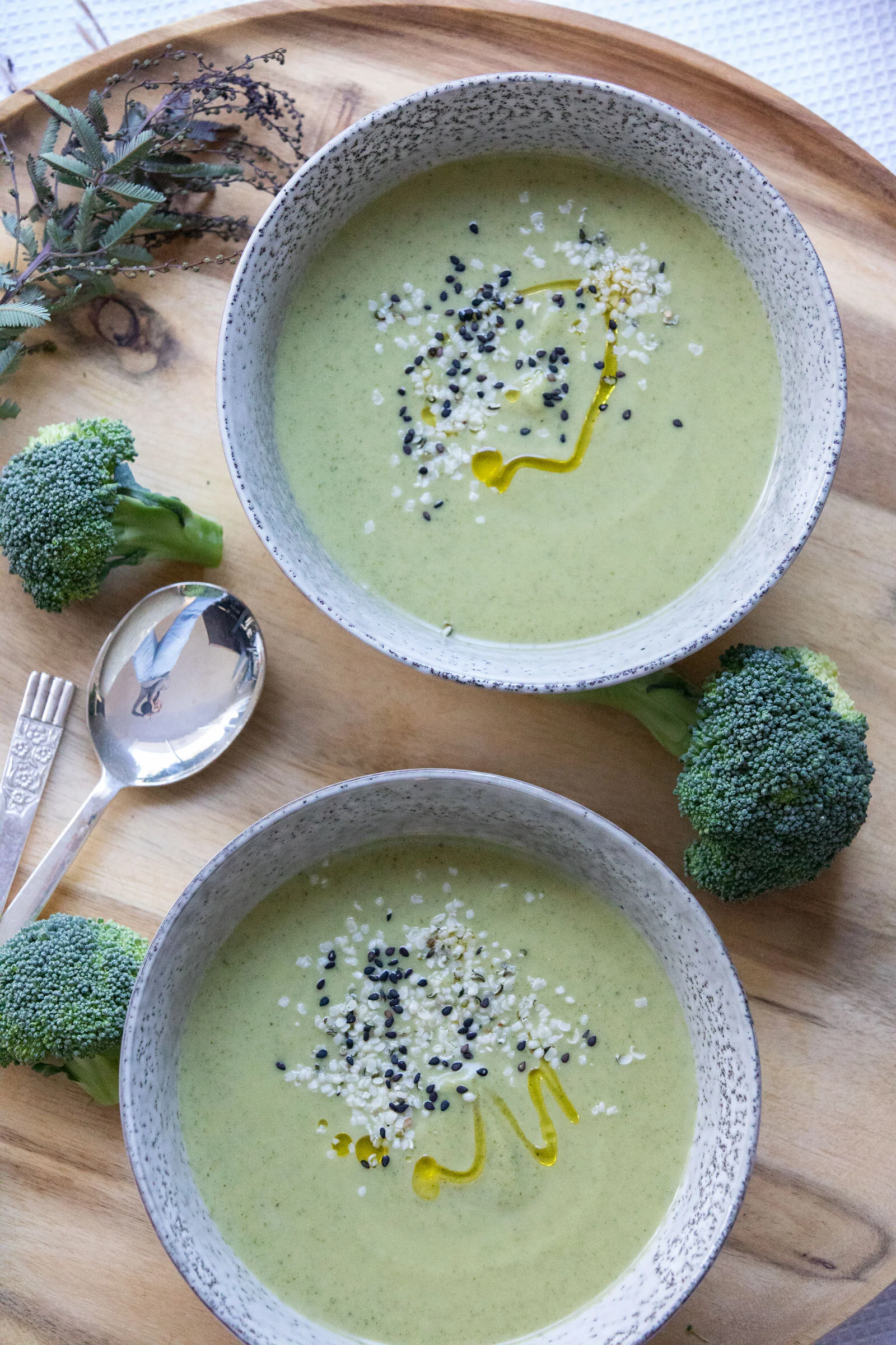 A bowl of green creamy soup garnished with black sesame seeds, white sesame seeds, a drizzle of olive oil, and a small amount of grated cheese. The bowl is placed on a wooden tray with broccoli florets and leafy herbs nearby.