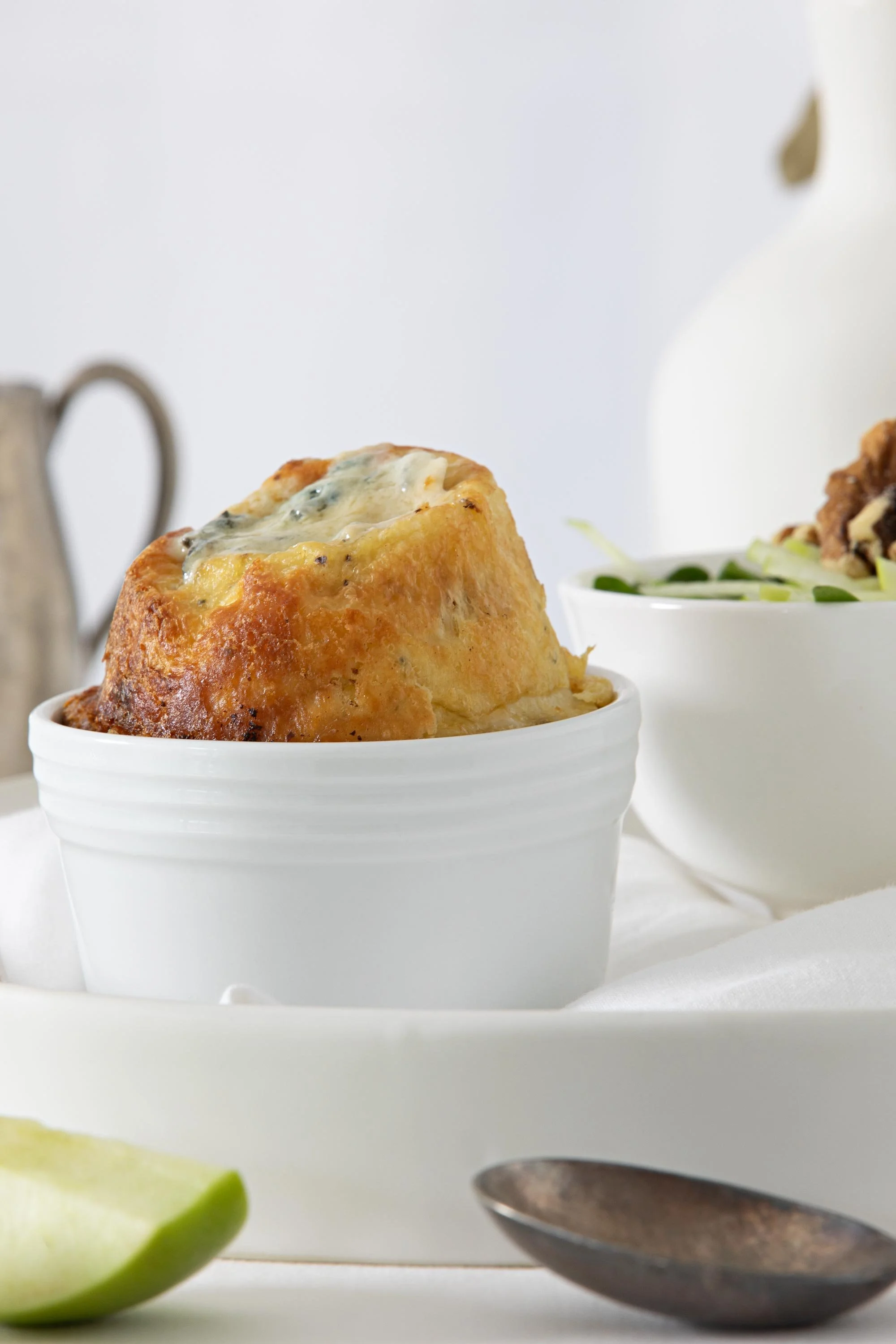 Close-up of a baked good topped with cheese and blue mold in a white ramekin, with a bowl of salad containing greens and fried chicken in the background, a piece of lime in the foreground, and a spoon partially visible at the bottom right.