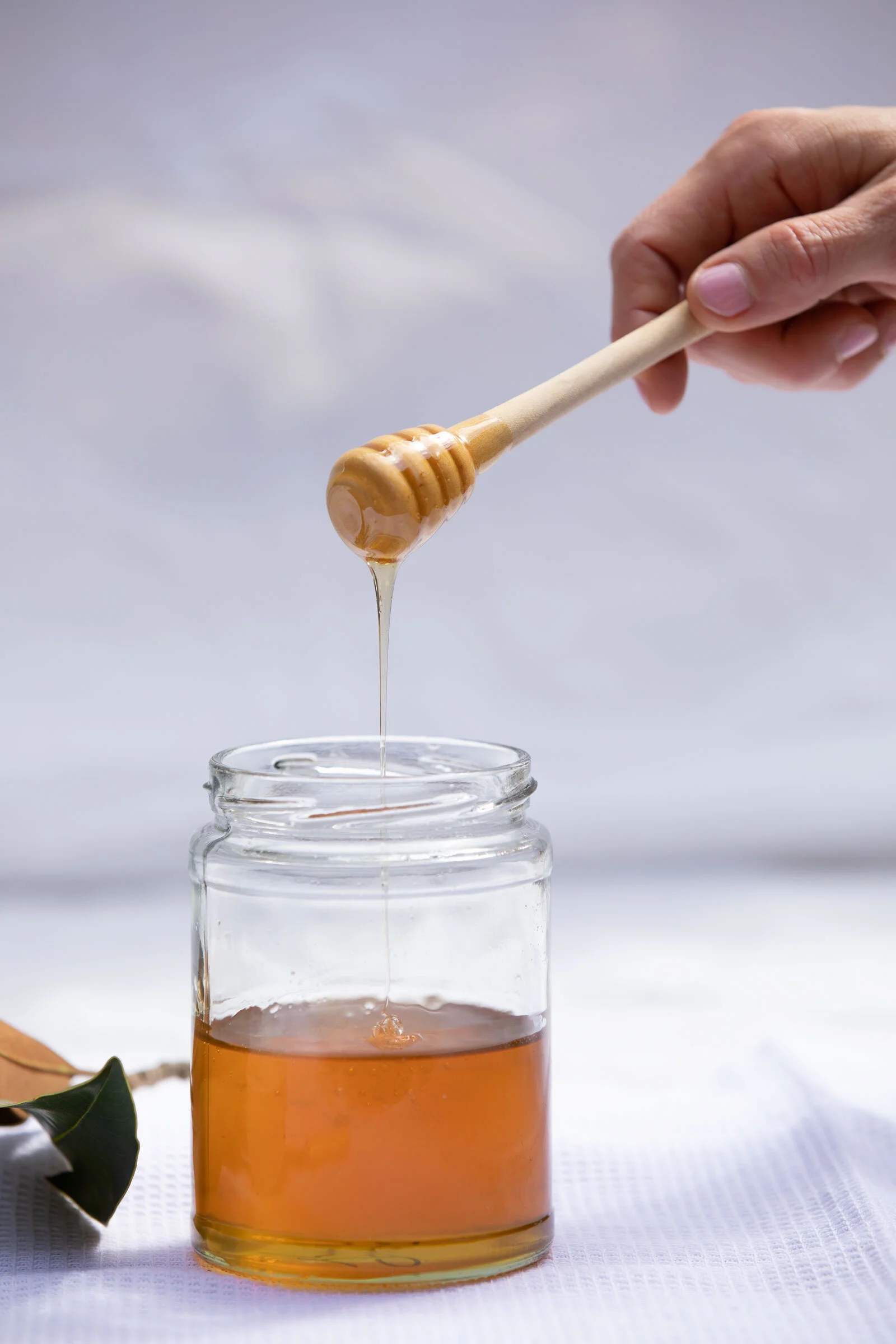 A hand holds a honey dipper dripping honey into a glass jar filled with honey, on a white surface with a blurred background.