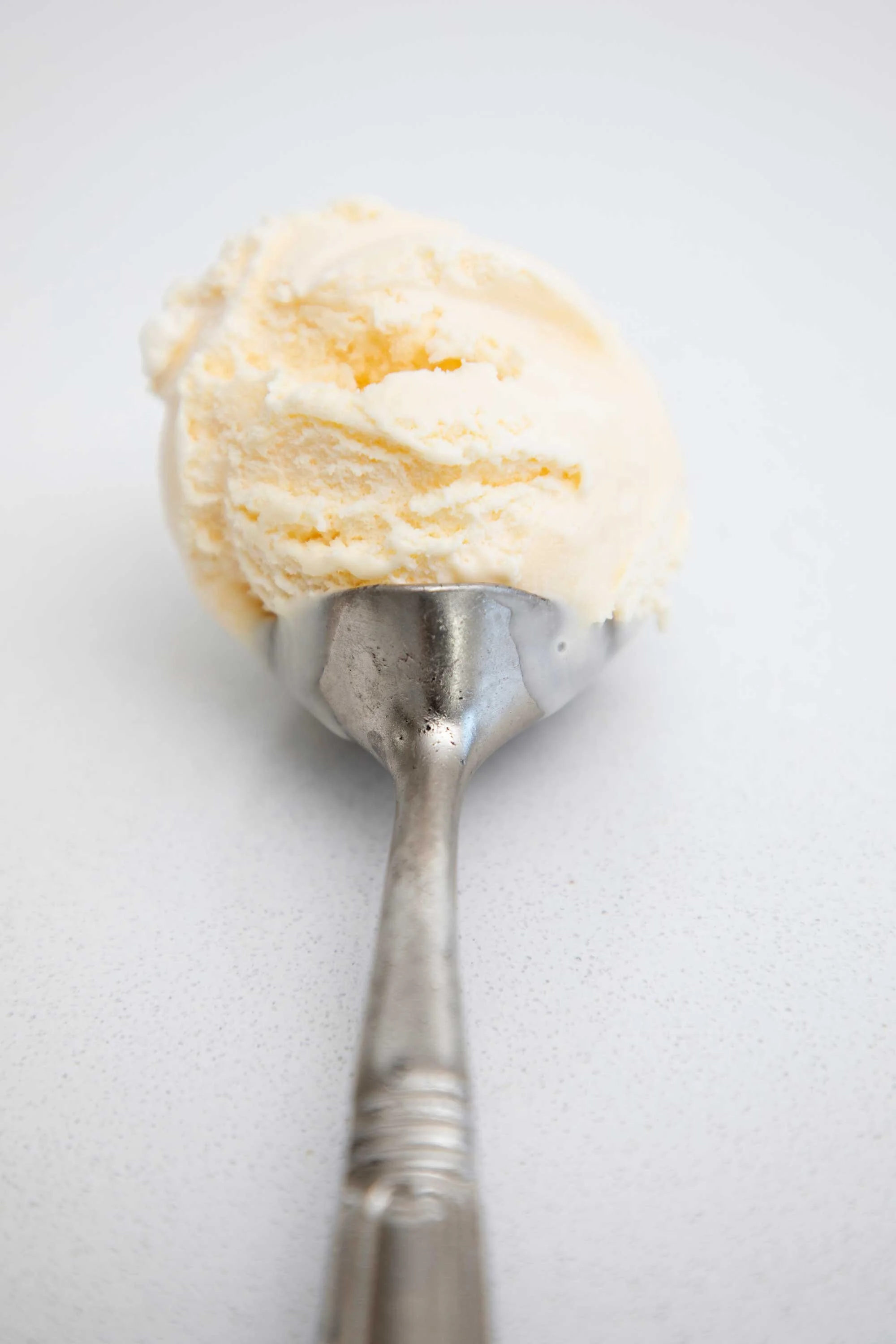A scoop of vanilla ice cream on a metal spoon. The background is a plain, light-colored surface.