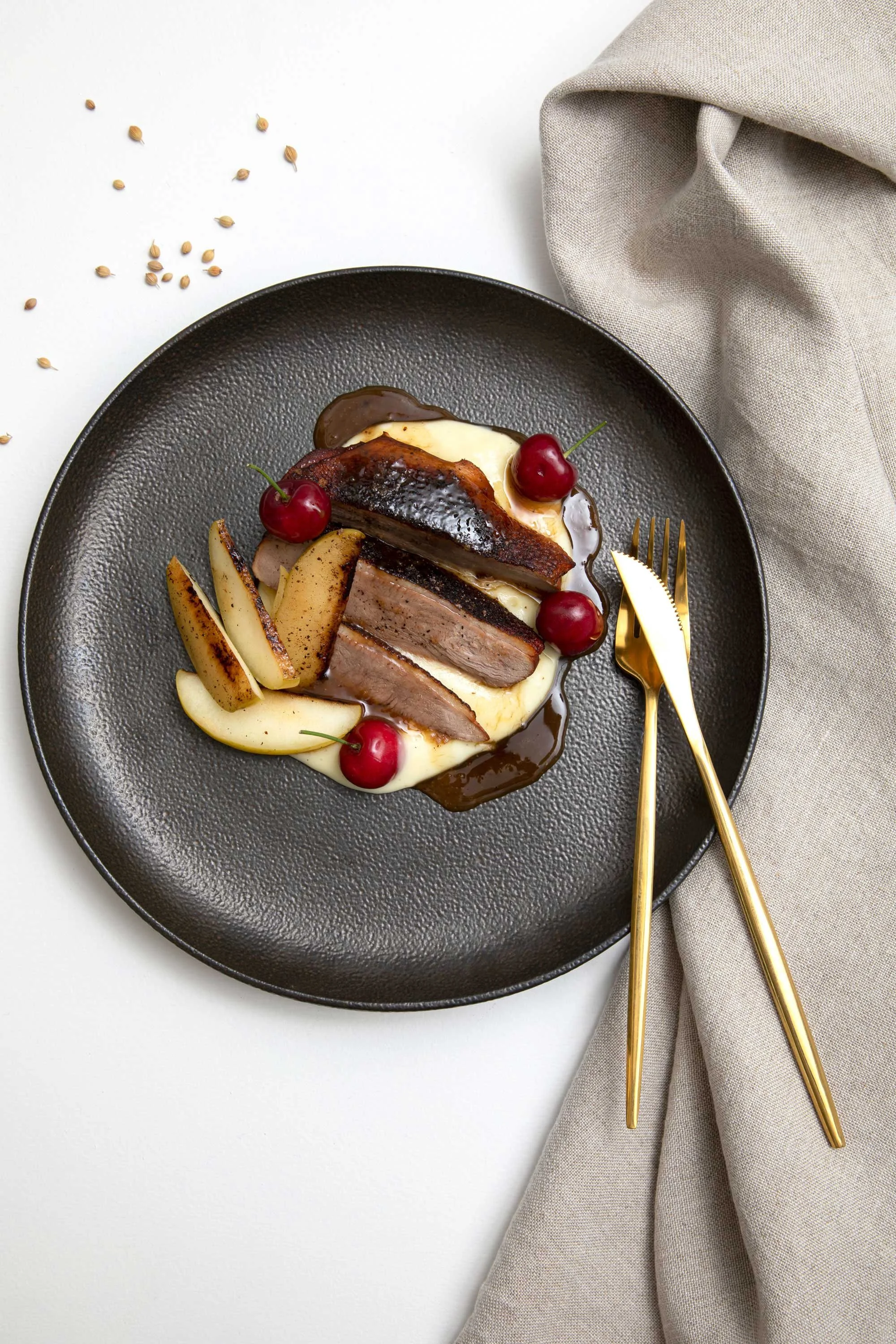 A black plate with slices of cooked meat, potato wedges, cherries, and a yellowish sauce, accompanied by a gold fork and knife, with a beige cloth napkin on the side.