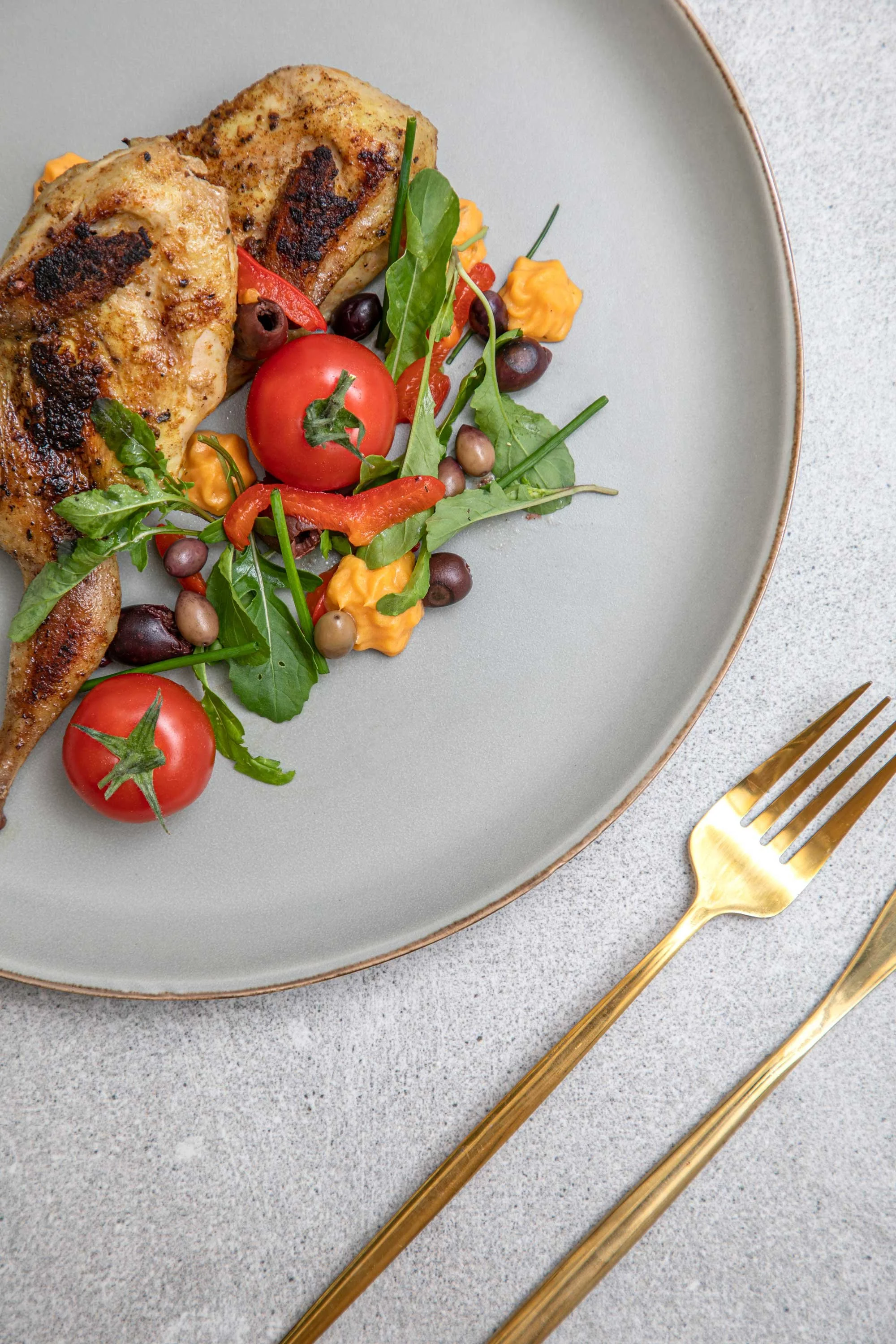 Plate with grilled chicken thighs and a side of cherry tomatoes, mixed greens, and small dollops of orange sauce, gold utensils placed beside on a textured grey surface.