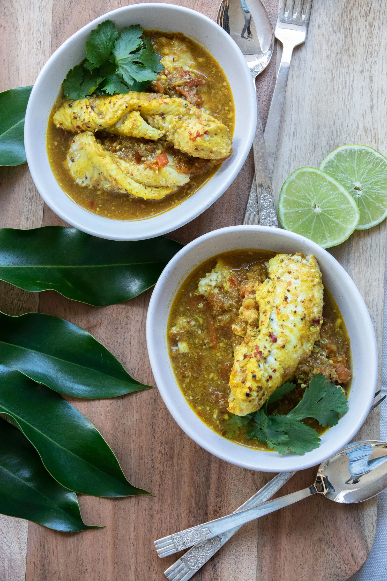 Two bowls of Indian fish curry garnished with cilantro, placed on a wooden table with lime wedges, metal cutlery, and green leaves for decoration.