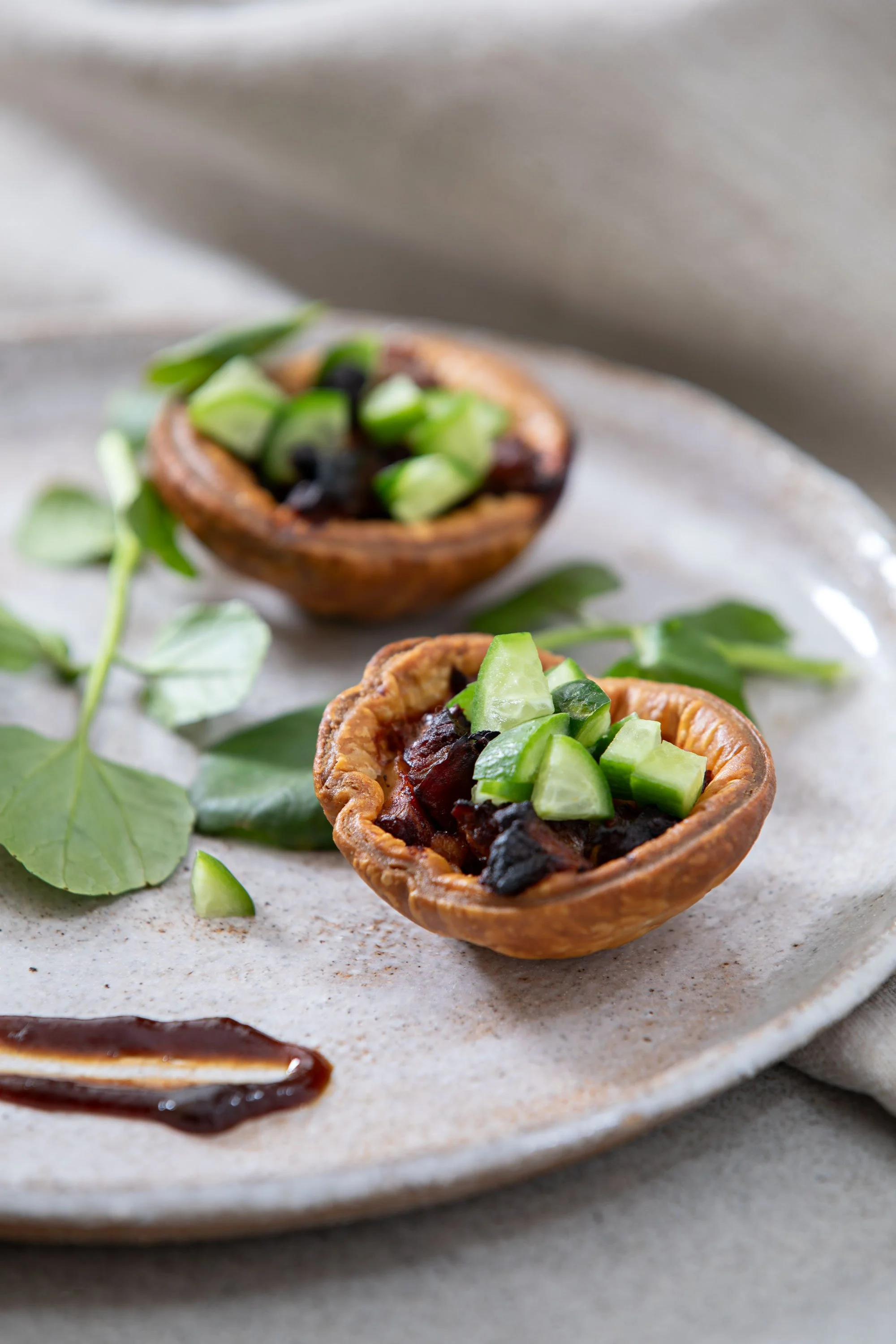 Two small savory pies filled with dark ingredients and topped with chopped green vegetables, placed on a ceramic plate with green leaves and a small pool of sauce drizzled nearby.
