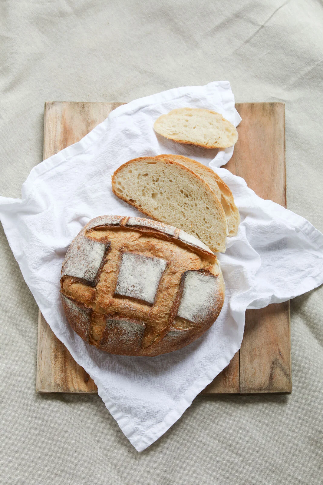A round loaf of rustic bread with a decorative pattern on top, two slices of bread, all placed on white cloth on a wooden surface.
