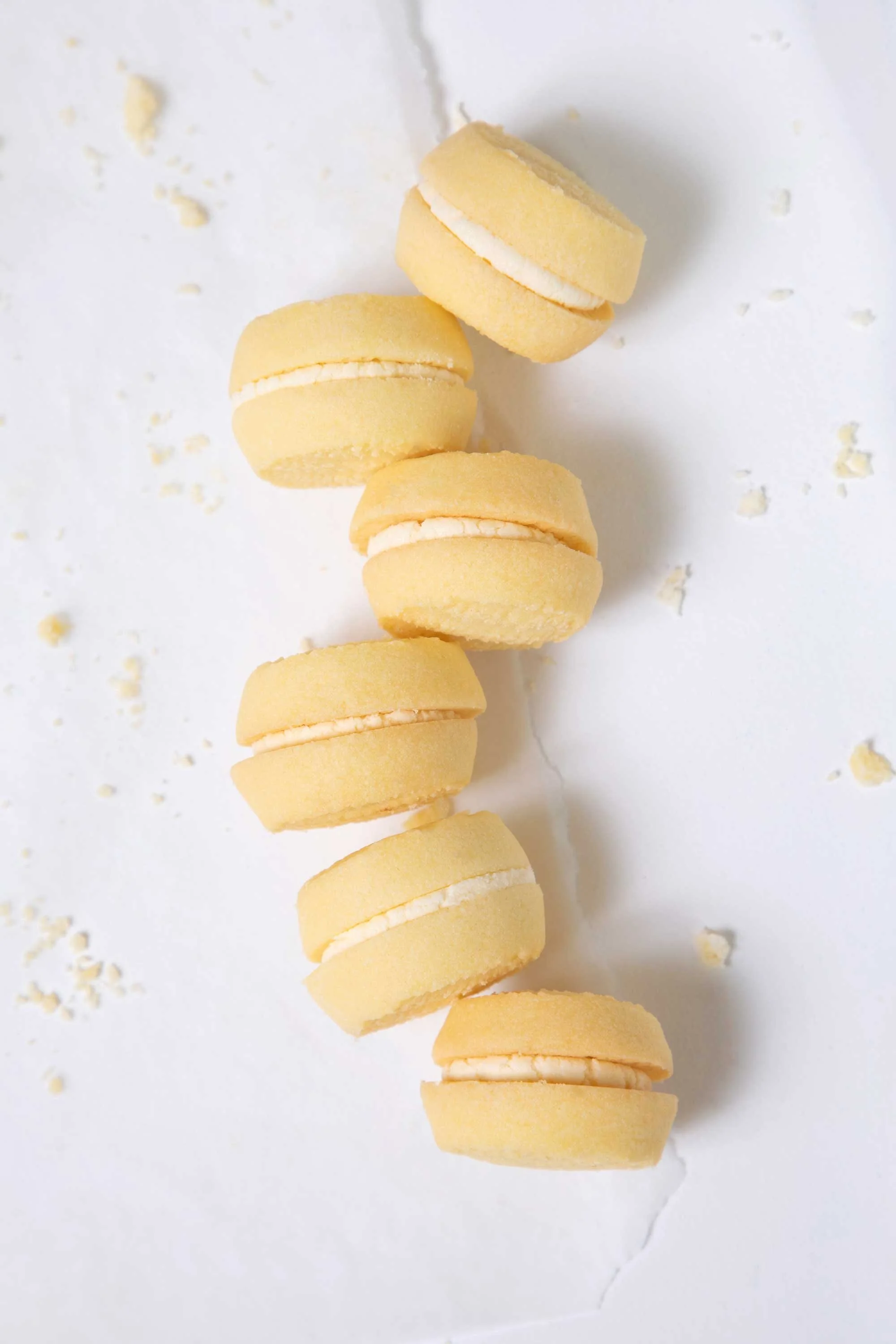 Line of yellow cookies with white filling on a white surface with cookie crumbs scattered around.