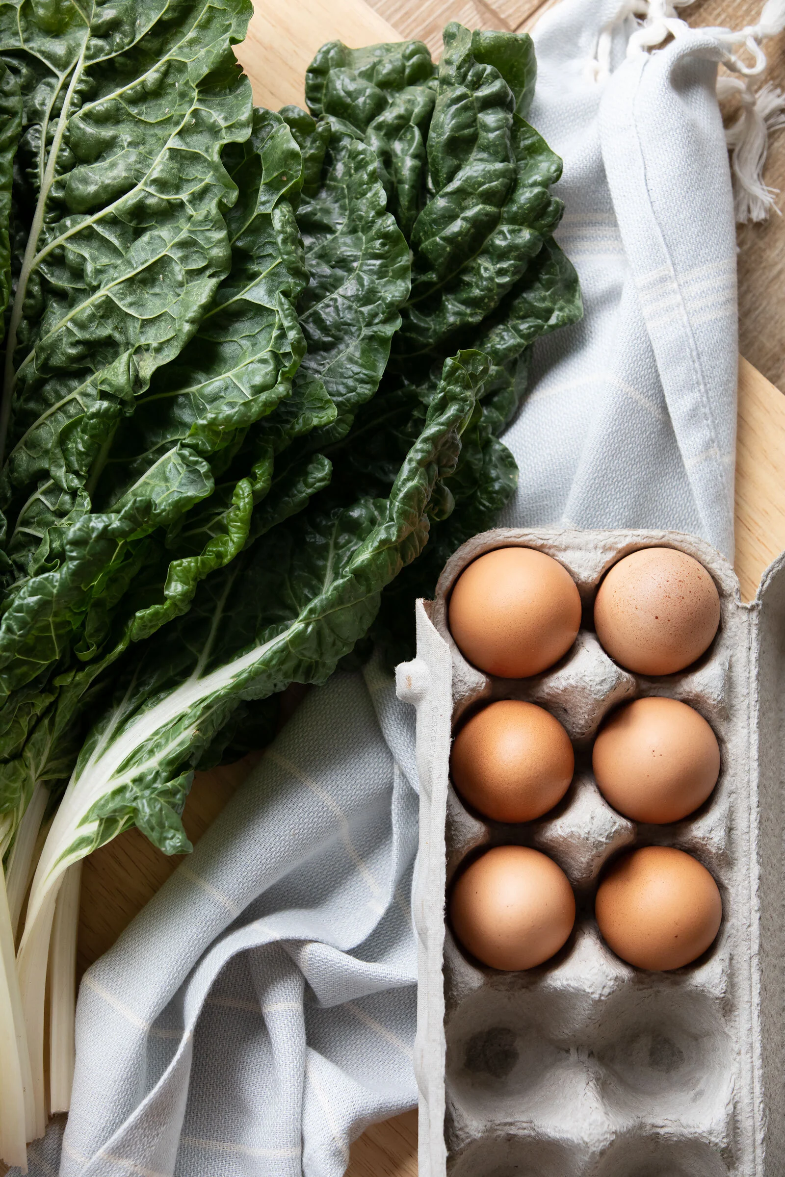 Fresh kale leaves on a wooden surface next to a carton of brown eggs.