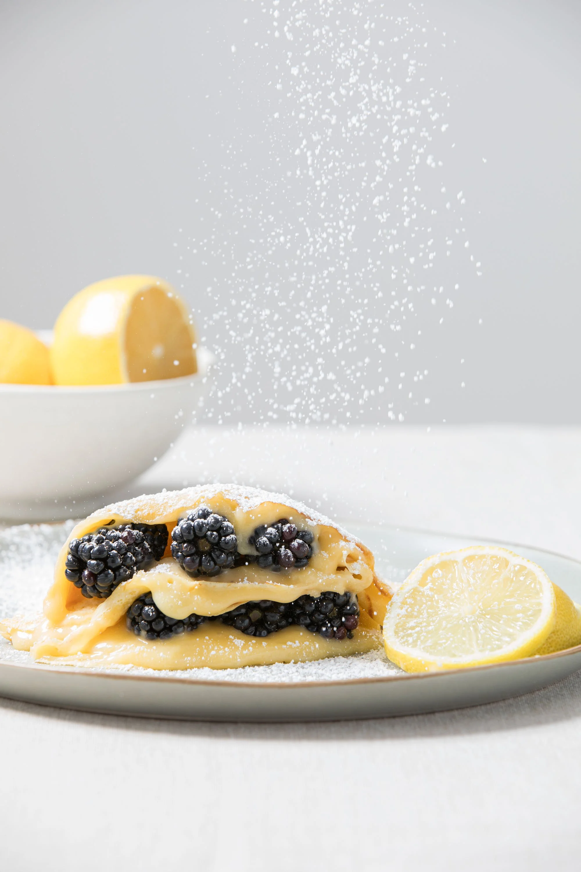 A plate with a dessert topped with blackberries and lemon slices, powdered sugar falling from above, and a bowl of lemons in the background.