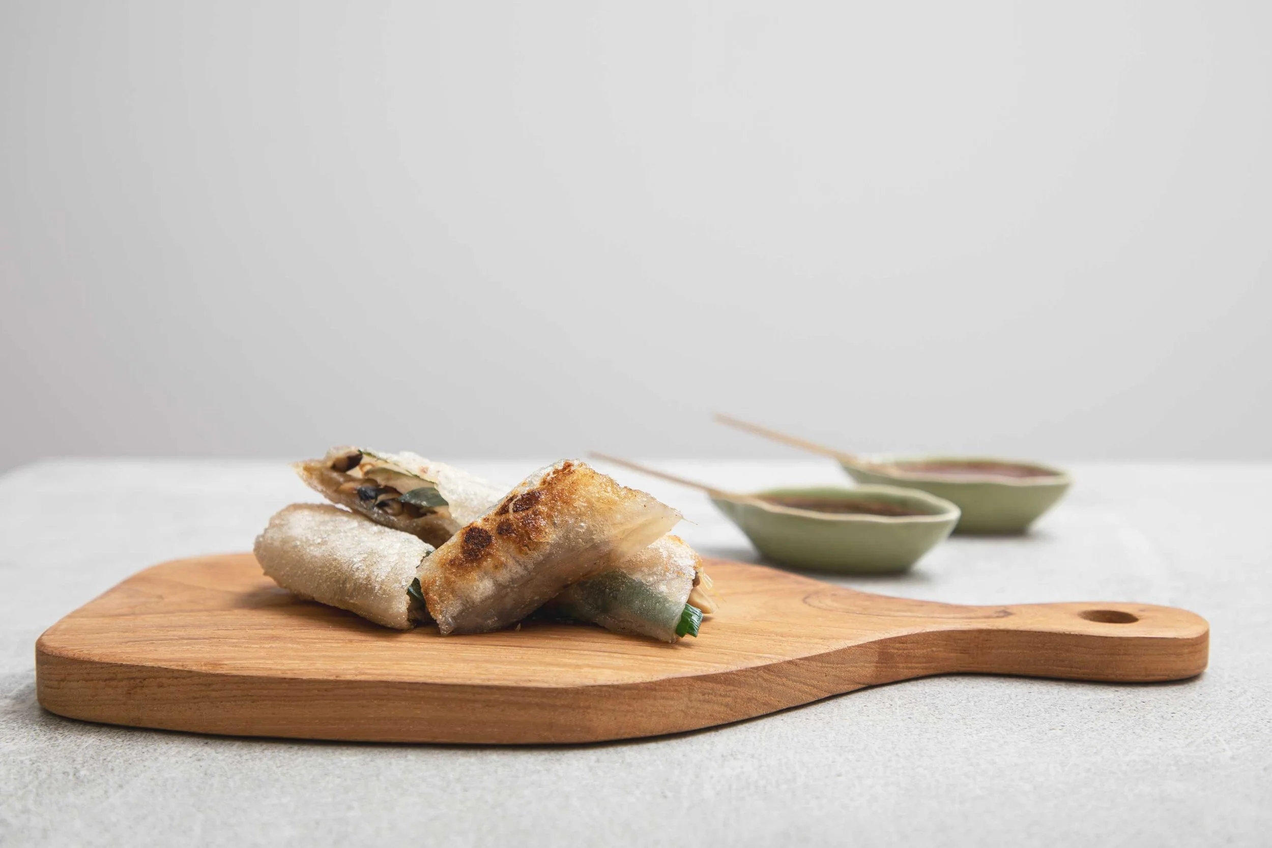 Stacked spring rolls on a wooden serving board with small dipping bowls in the background.