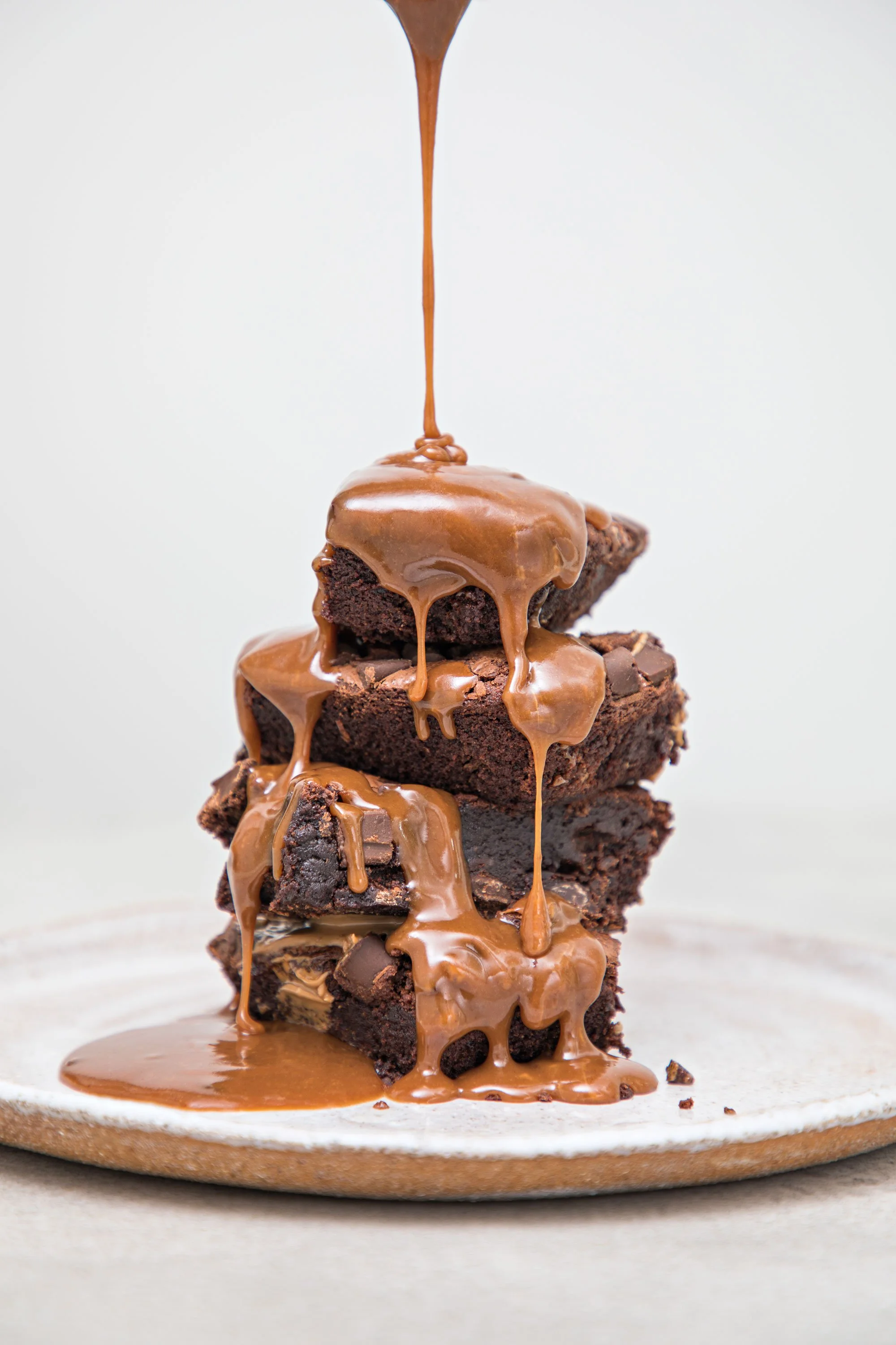 Stack of four chocolate brownies topped with melted caramel which is dripping down the sides, on a white surface with caramel spilled around.