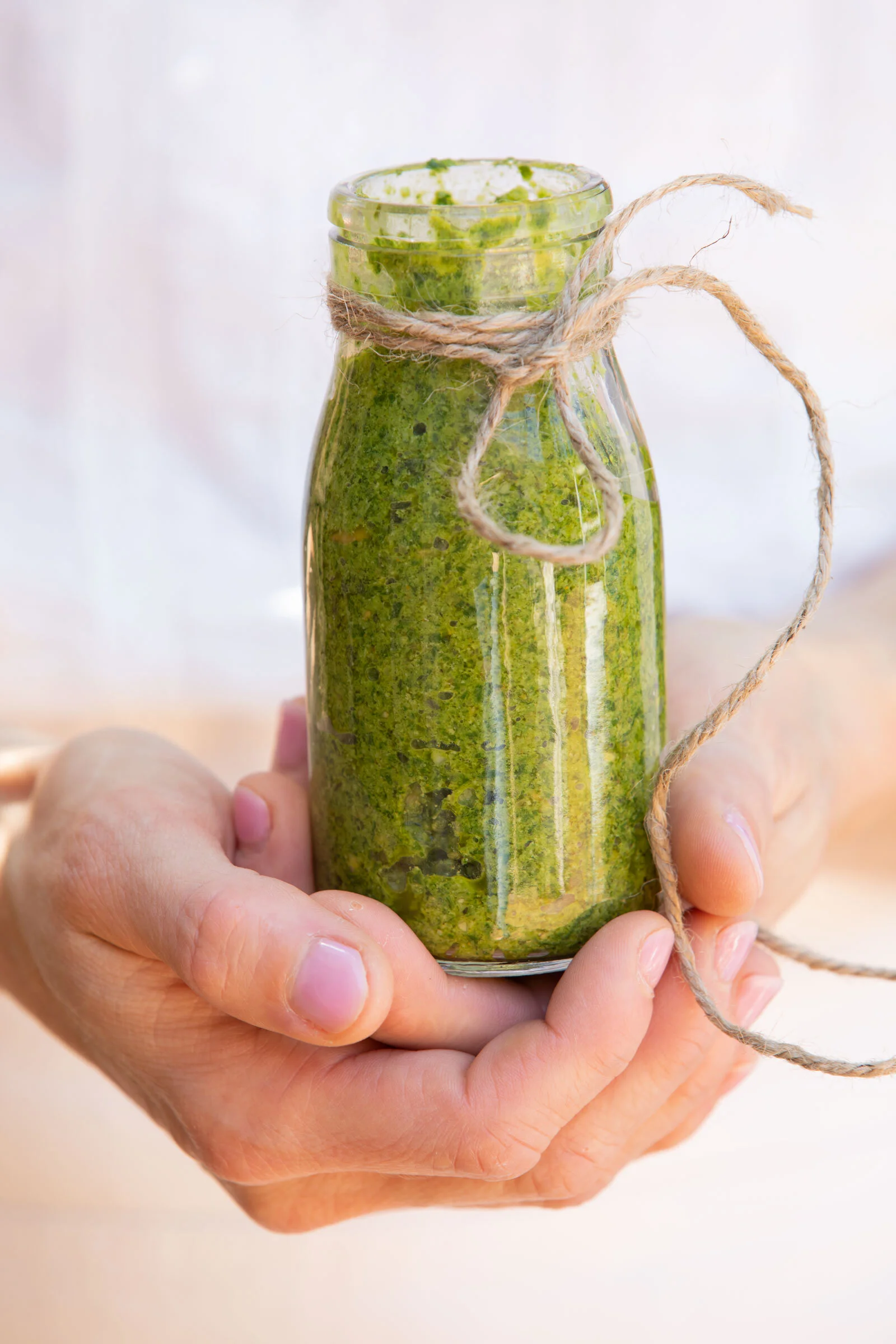 A person holding a small glass jar filled with a green smoothie, with a piece of twine tied around the neck of the jar.