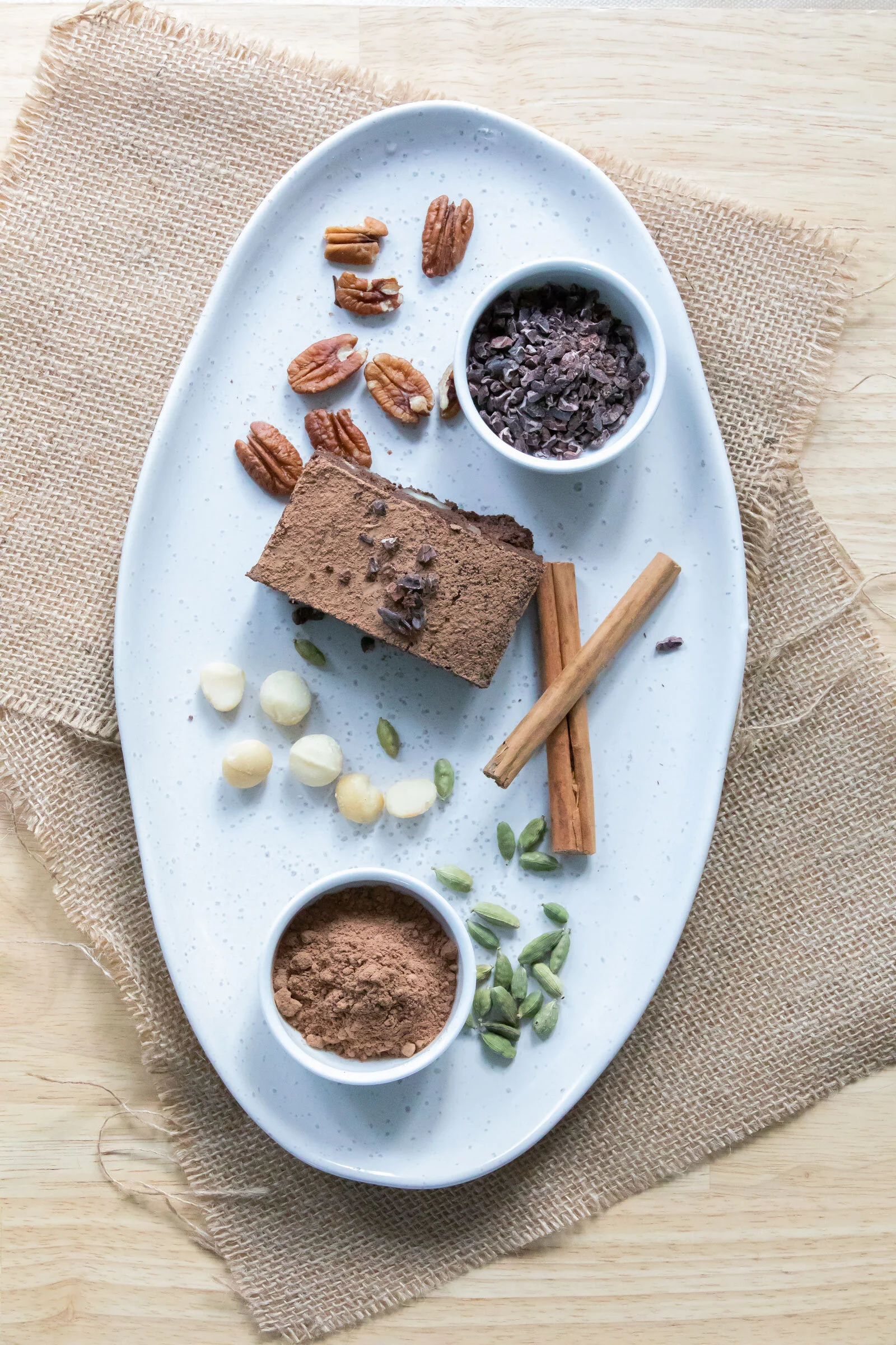 A white oval plate with a piece of chocolate cake, a small bowl of chocolate shavings, pecans, cinnamon sticks, white chocolate chips, and green pumpkin seeds, on a wooden surface with a burlap cloth underneath.