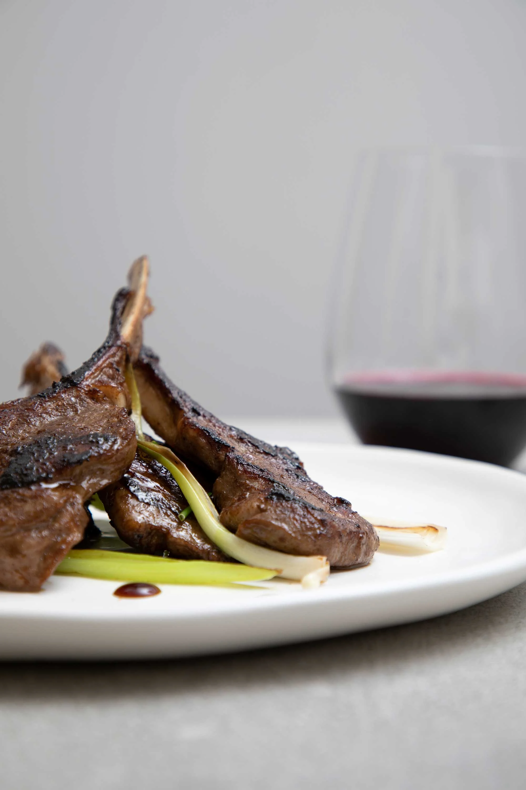 Close-up of grilled lamb ribs garnished with greens on a white plate, with a glass of red wine in the background.