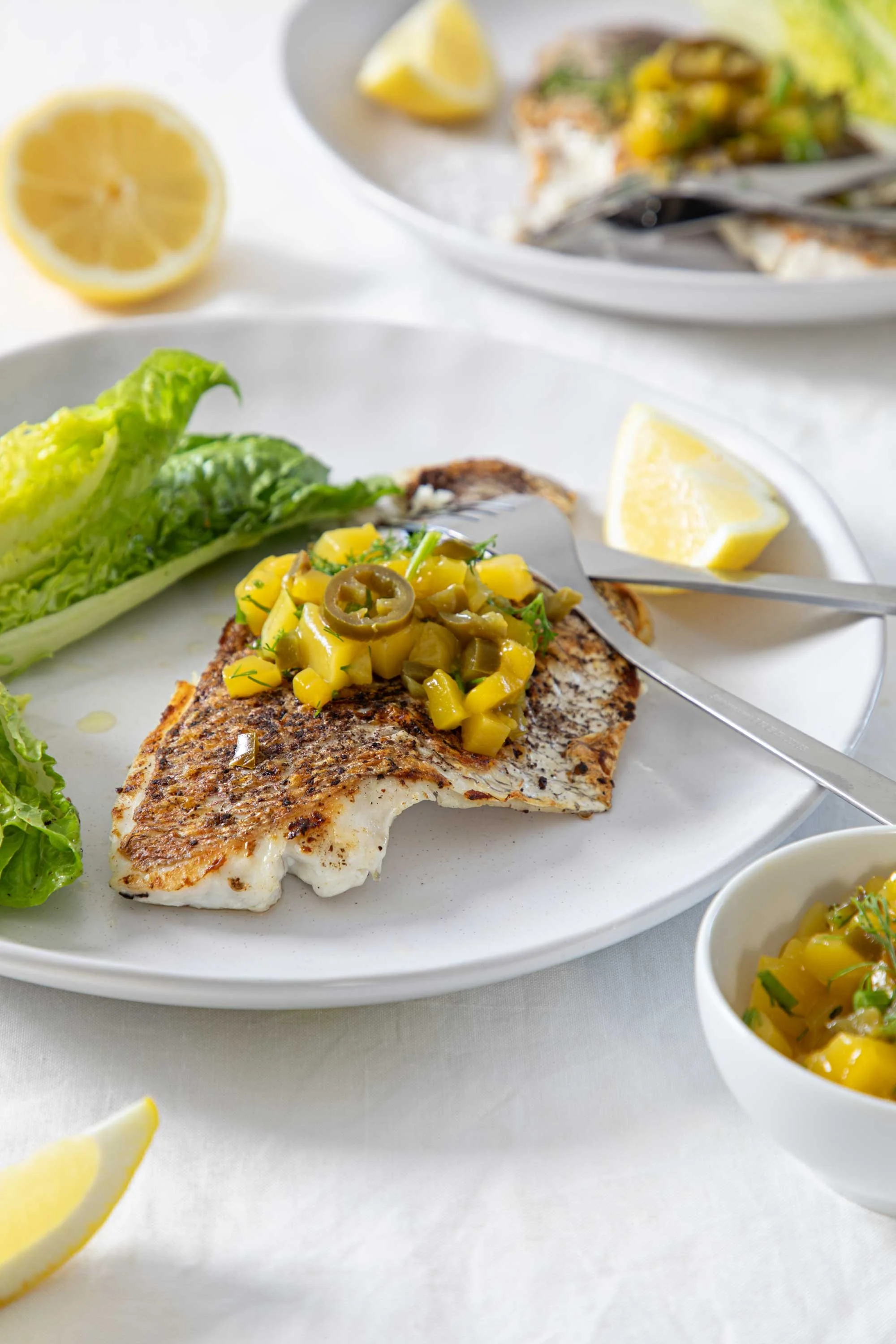 Grilled fish fillet topped with mango and jalapeño salsa on a white plate, with a side of lettuce and lemon wedges, and a small bowl of mango salsa.