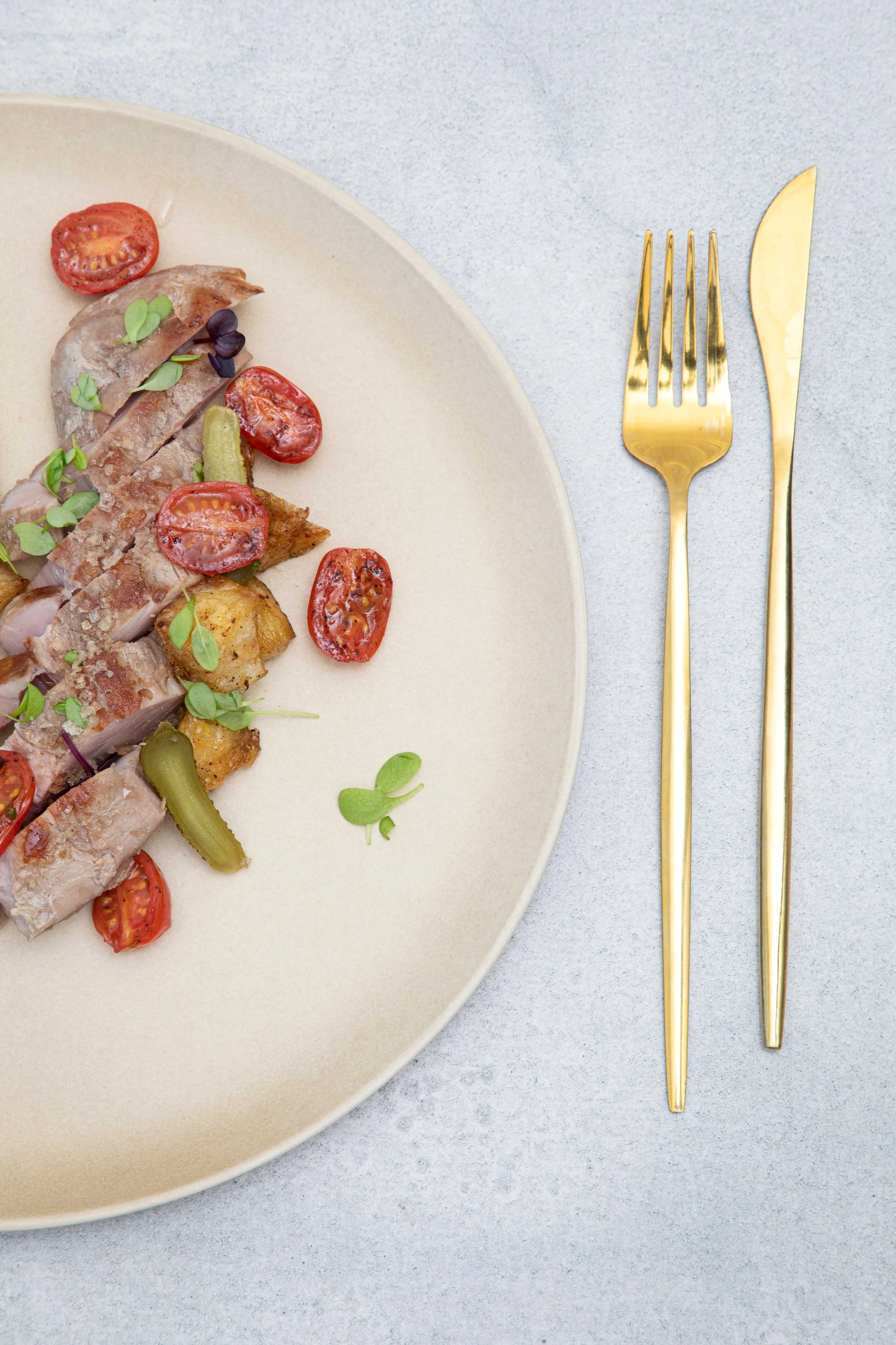 A beige plate with sliced grilled chicken topped with cherry tomatoes, microgreens, olives, and pickles, placed on a light gray surface with a gold fork and knife beside it.
