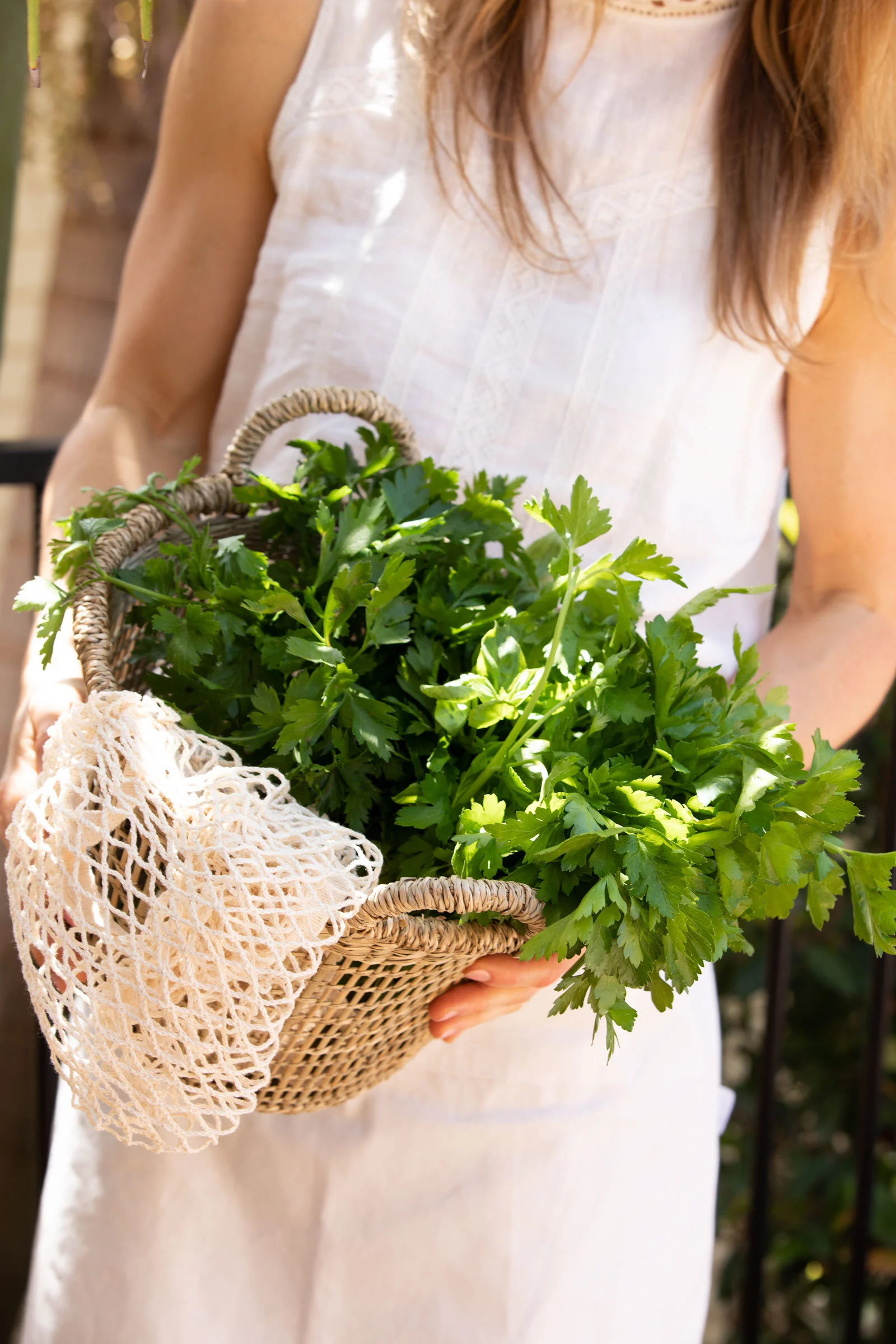 Person in a white dress holding a basket of fresh green herbs, with a cream wool bag hanging from the basket's side.