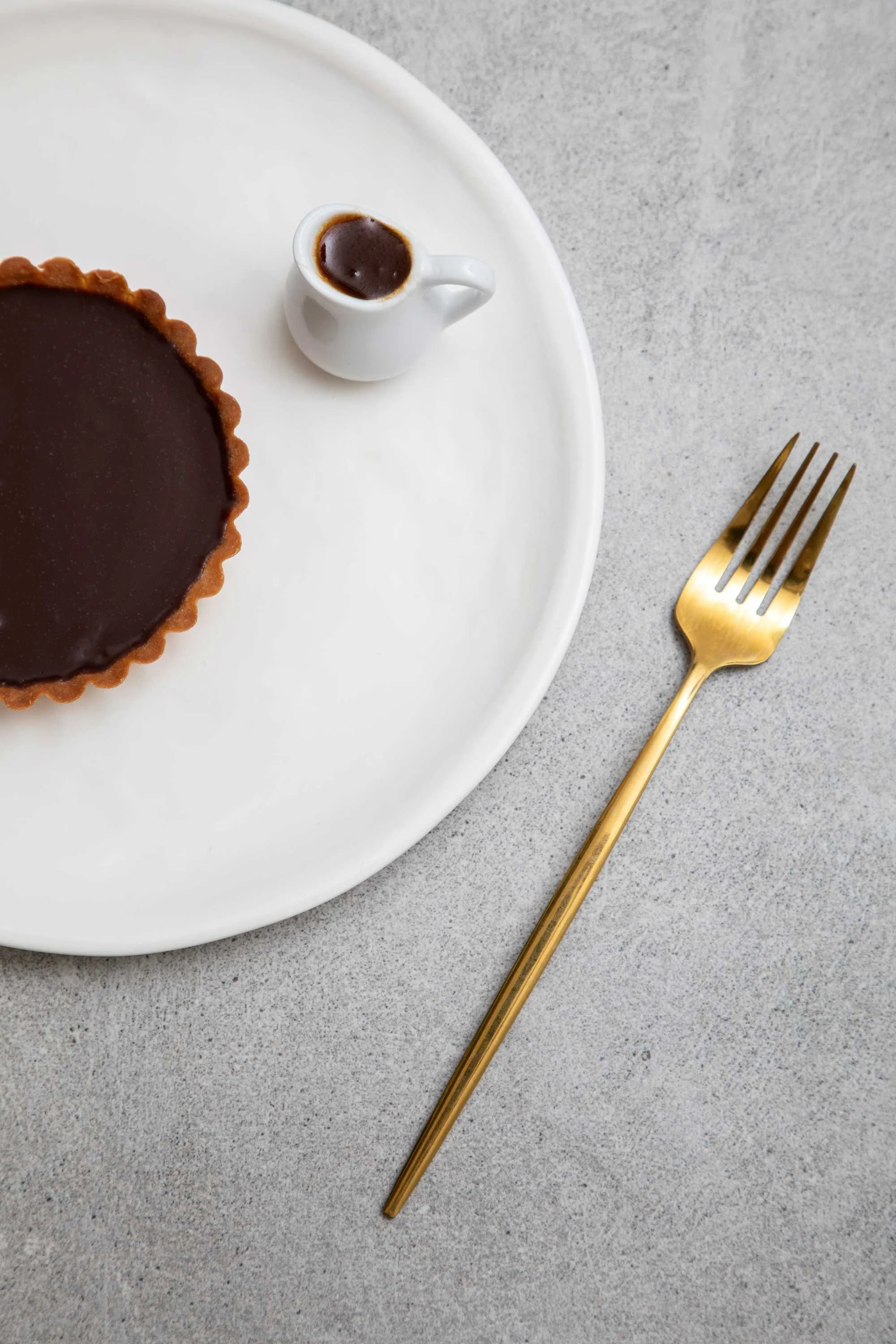 A white plate with a chocolate tart, a small white pitcher filled with chocolate sauce, a gold fork, and a gray surface in the background.