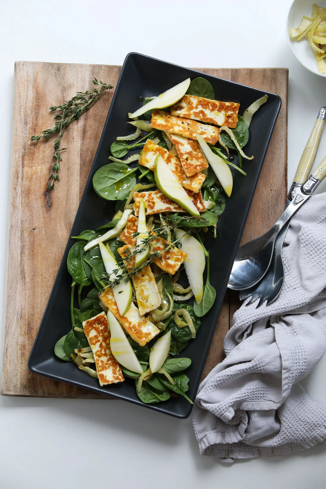 Fresh salad with greens, sliced pears, grilled halloumi cheese, garnished with thyme on a black rectangular plate, on a wooden board with a spoon wrapped in a white cloth and a small dish of pasta in the background.