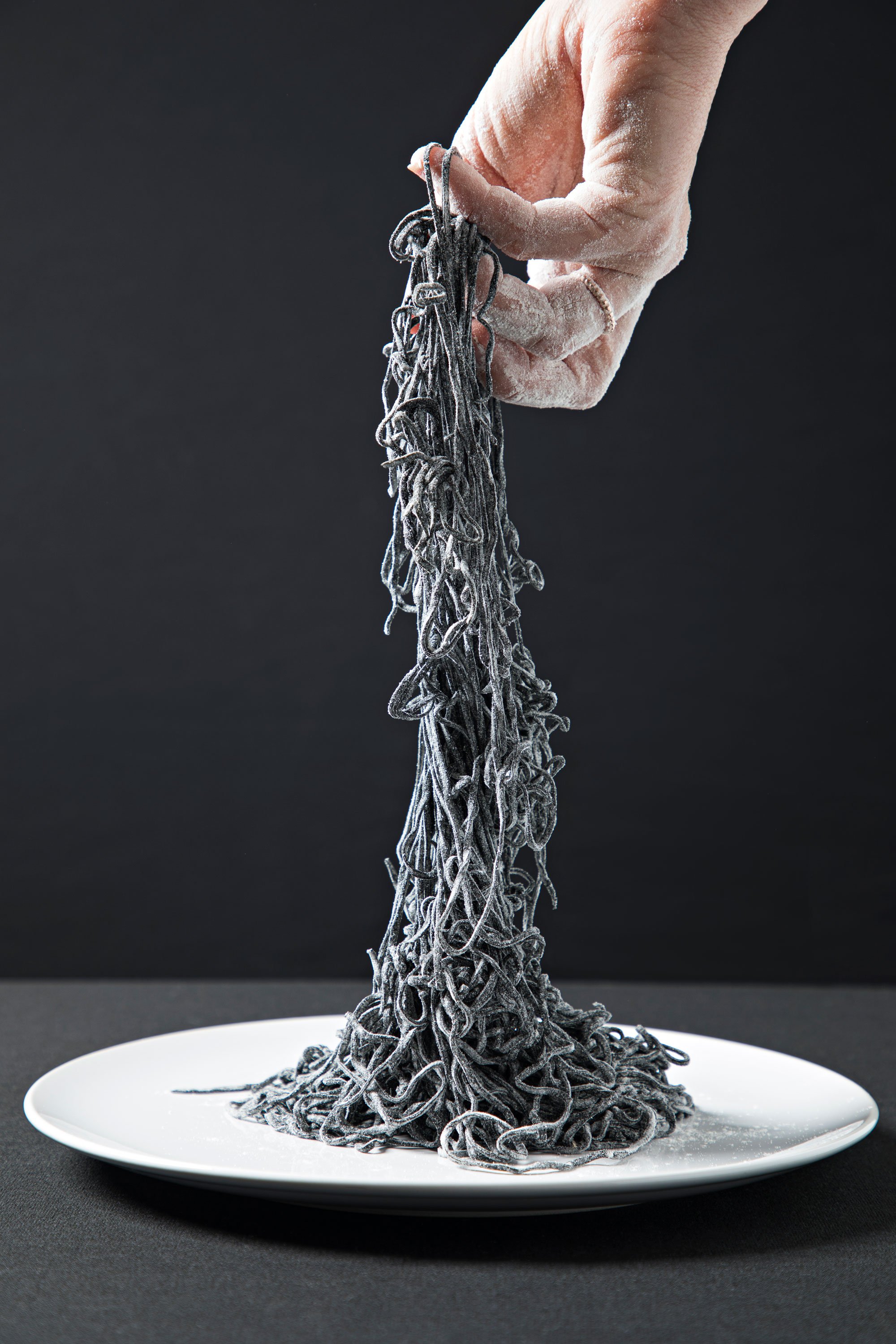 A white plate holds a tall, narrow mound of black, tangled spaghetti with some strands spilling over the edge onto the plate, set against a black background.