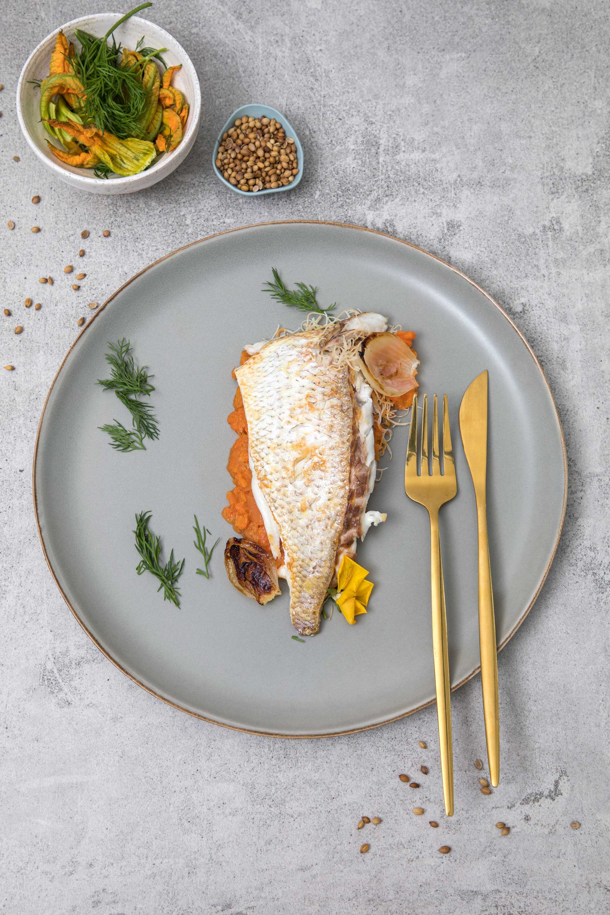 A plate with a cooked fish fillet on a bed of orange vegetables, garnished with fresh dill, and accompanied by a yellow edible flower. A fork and knife with gold handles are placed next to the dish. In the background, a small bowl of green herbs and 