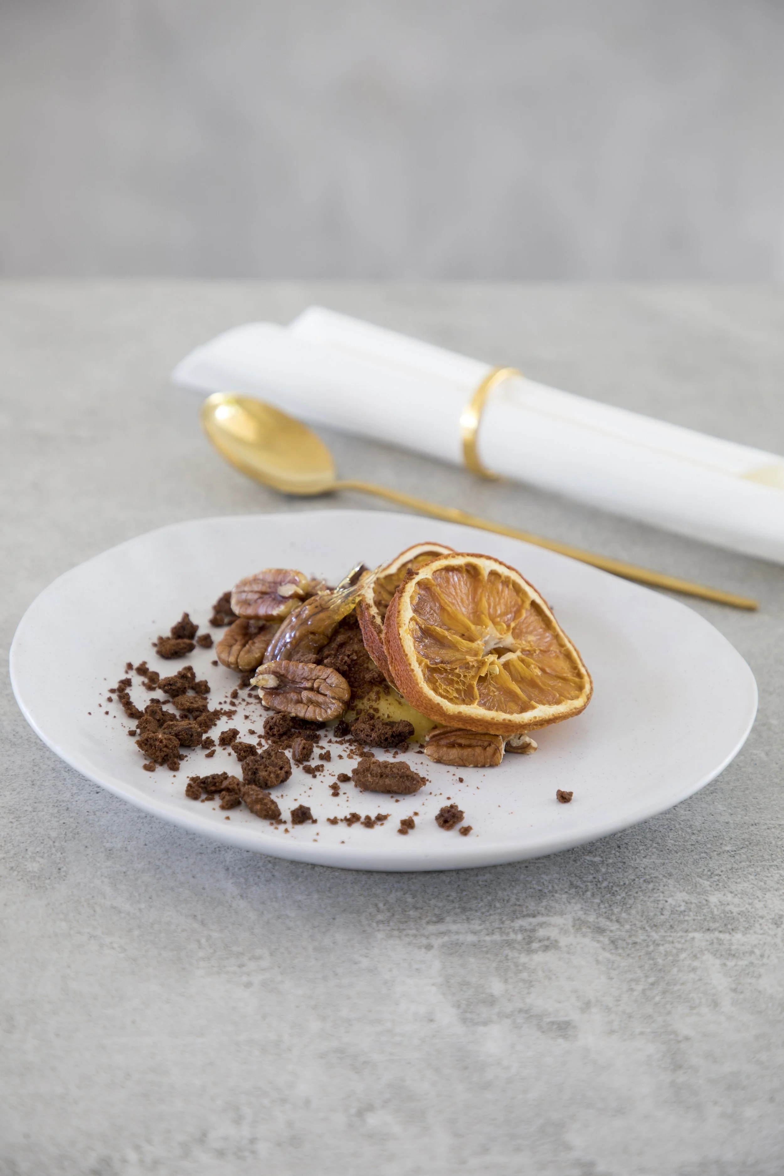 Dessert plate with chocolate cake, pecans, dried orange slices, surrounded by chocolate crumbs, on a gray surface with gold cutlery and a white napkin.