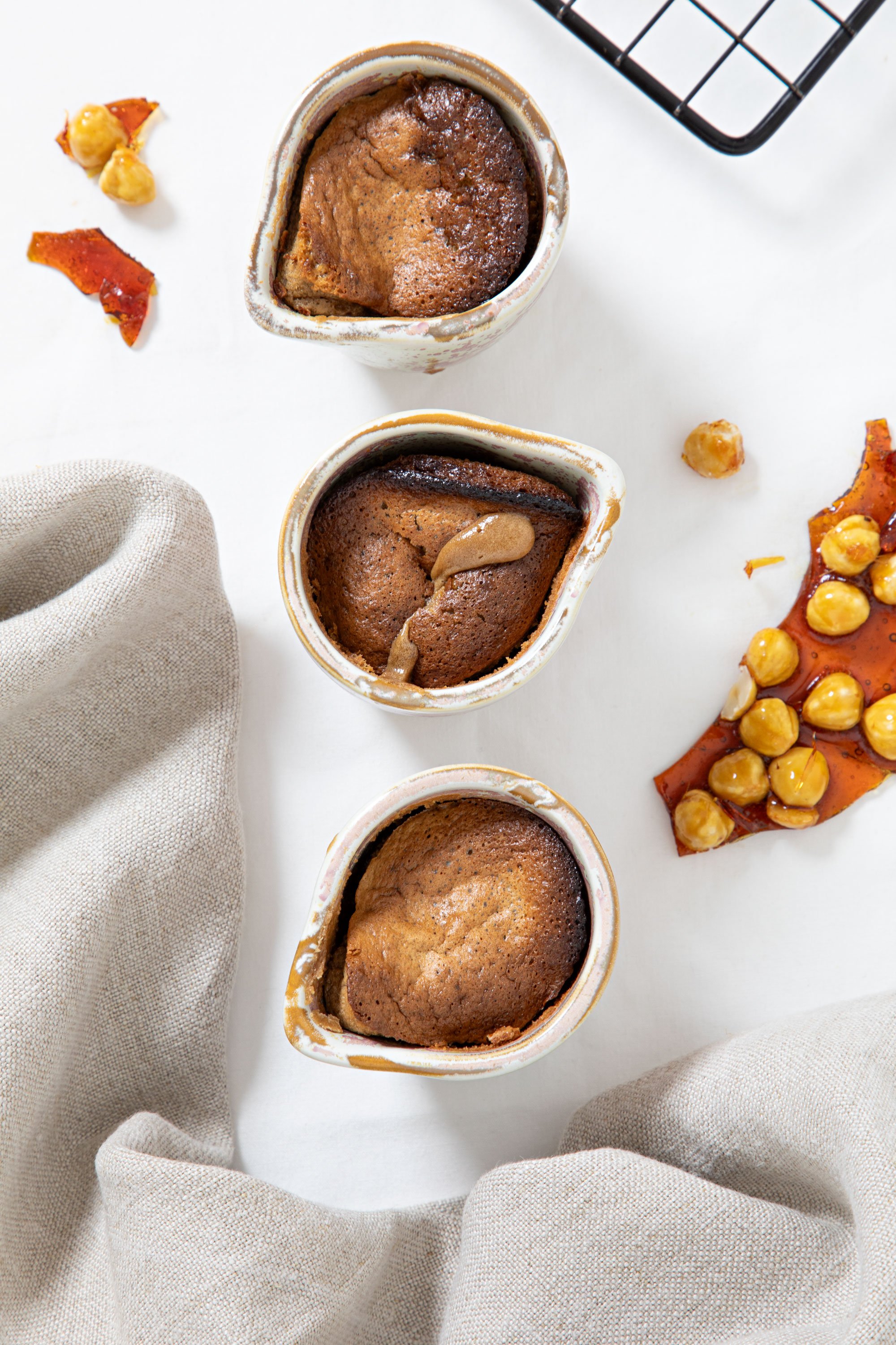Three ramekins of baked chocolate desserts on a white surface, with a beige cloth and a baking rack nearby, garnished with caramelized nuts and slices of candied fruit.