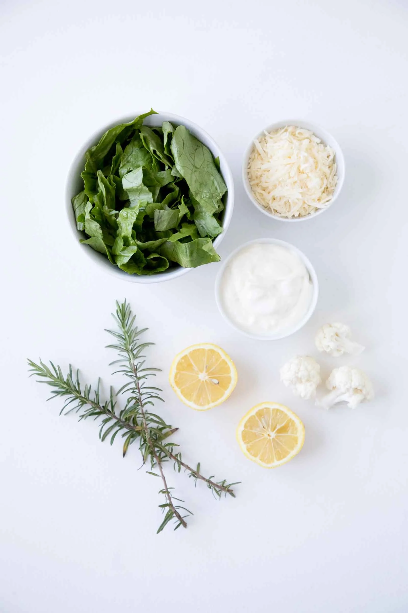A white surface with bowls of leafy greens, shredded cheese, and a white sauce, along with slices of lemon, cauliflower, sprigs of rosemary, and a piece of cauliflower.