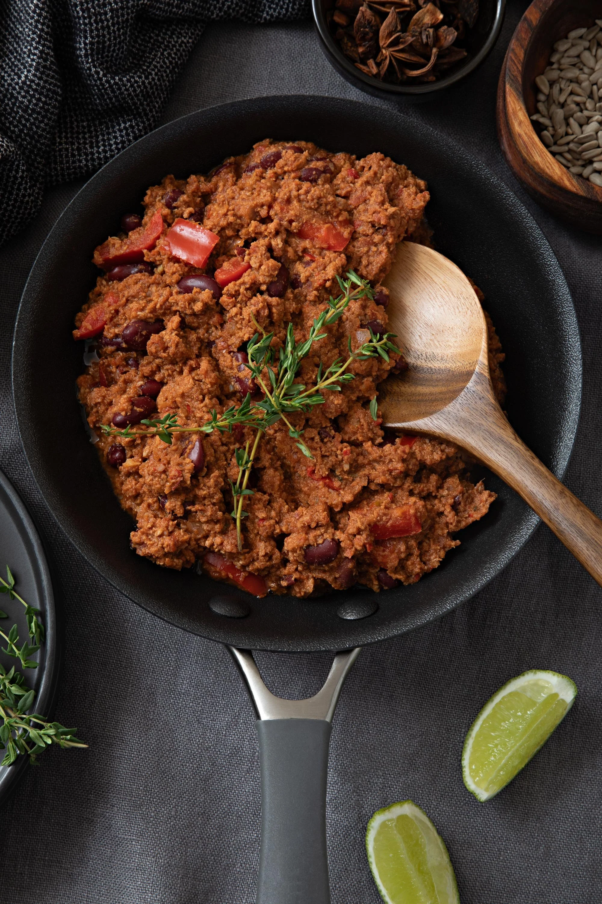 A cast iron skillet filled with cooked ground meat and beans, garnished with a sprig of fresh thyme, with lime wedges nearby, on a dark textured surface.